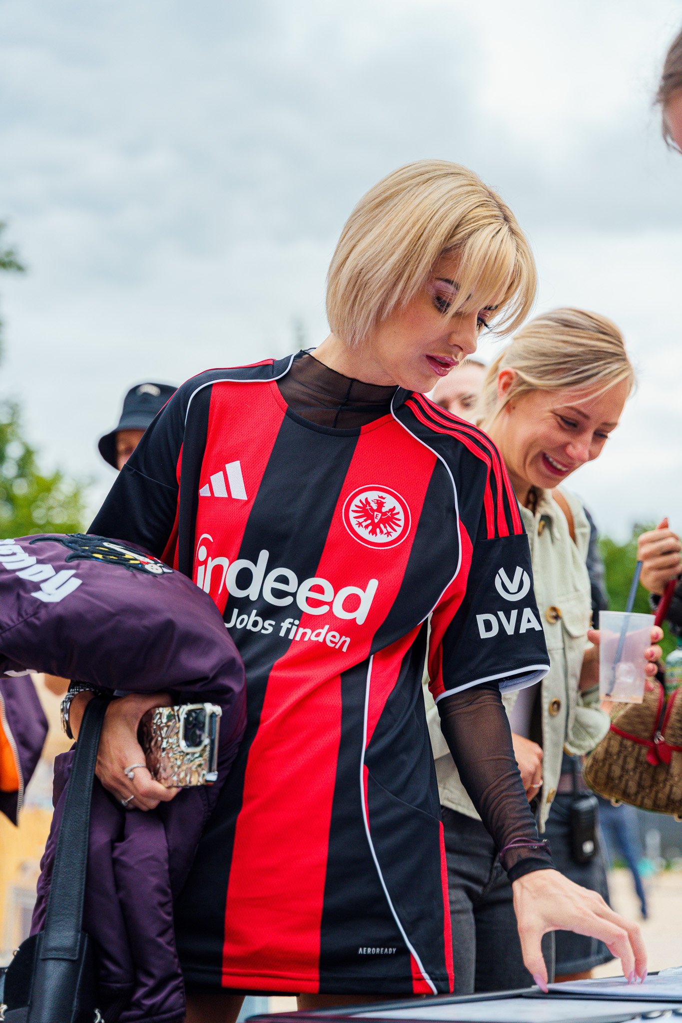 A woman in a red and black soccer jersey with logos, holding items and looking down, surrounded by other people outdoors.