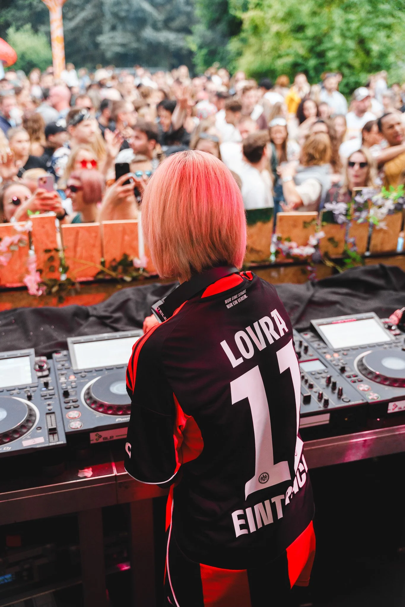 A female DJ wearing a black and red jersey with the name 'LOIRA' and the number 11 stands behind DJ equipment at an outdoor event, facing a large crowd of people.