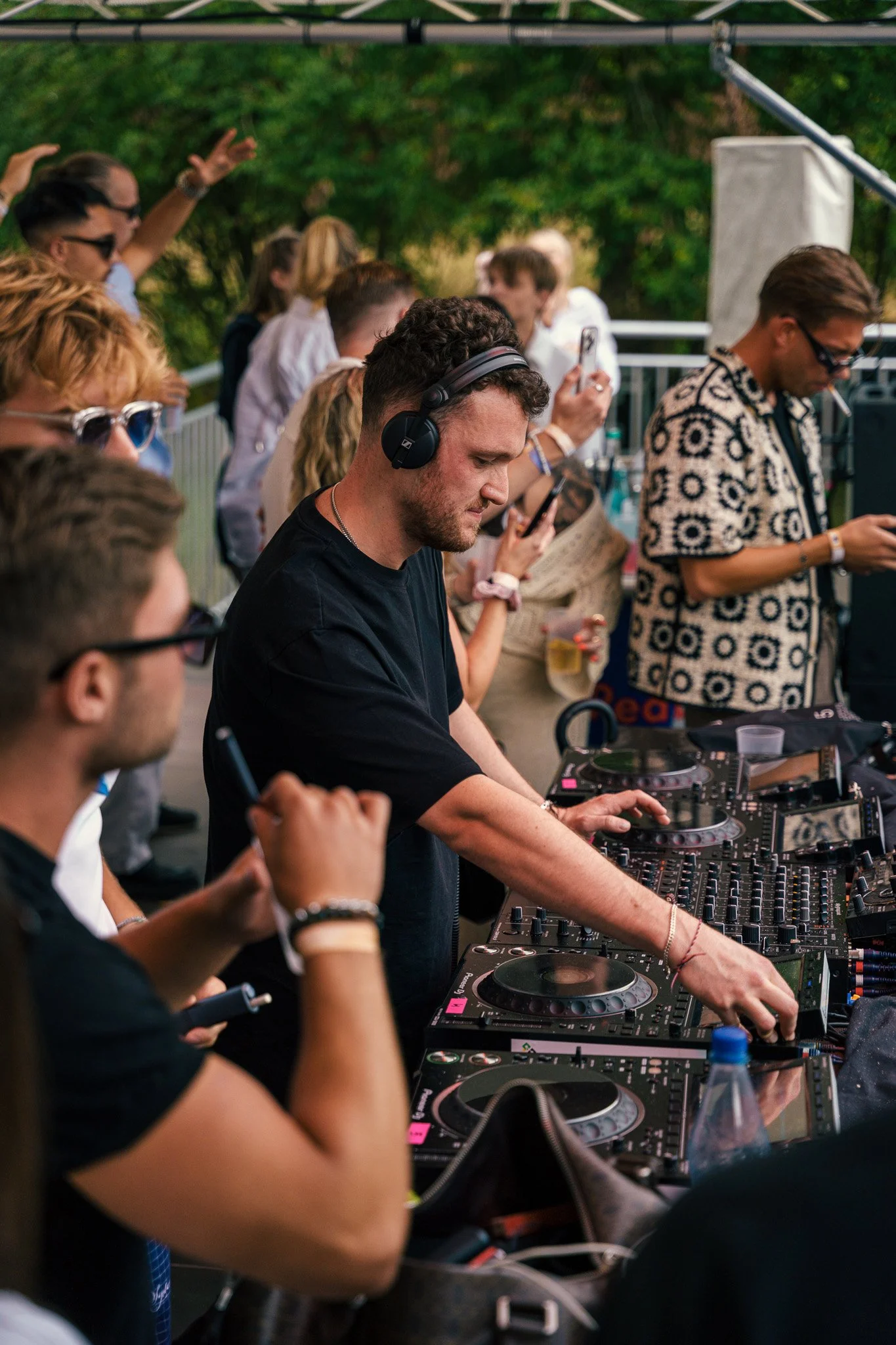 A young male DJ wearing headphones and a black shirt operates a DJ mixer at an outdoor event, surrounded by a crowd of people dancing and taking photos.