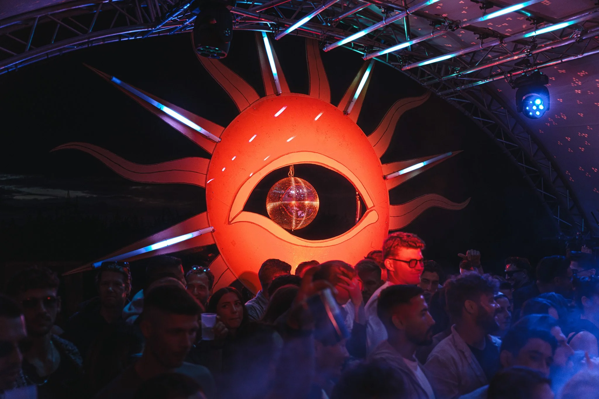 A crowd of people attending a music event with a large decorative sun and eye installation in the background, illuminated with colorful lights and a disco ball hanging in the sun's eye.