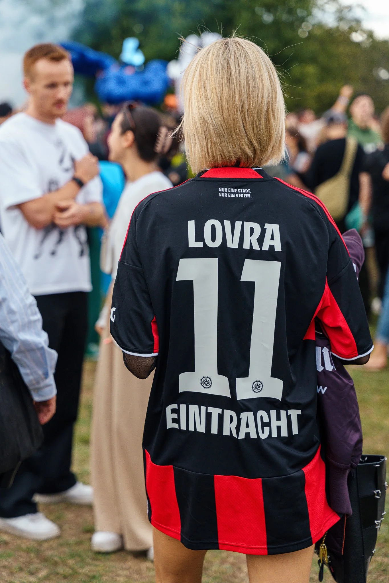 Back view of a woman wearing a black and red soccer jersey with the name 'LOVRA' and the number '11' on the back, at a crowded outdoor event.