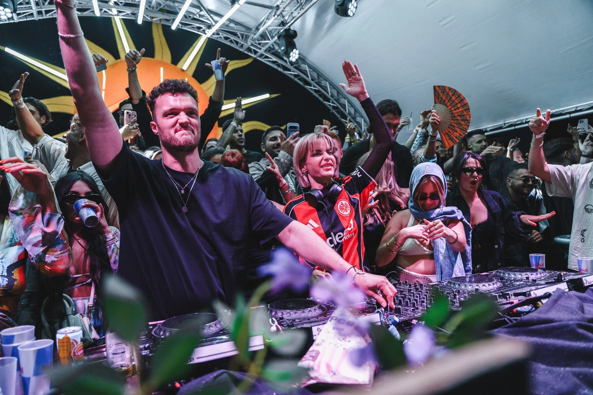 Crowd of people dancing and enjoying music at an indoor event with a DJ booth in the foreground and decorative lights overhead.
