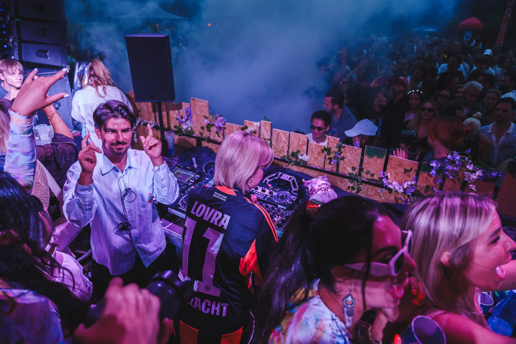 A DJ booth with a man and woman among a crowd at a lively event. The man is dancing and smiling, wearing a white shirt; the woman is working the DJ equipment, wearing a black jacket with 