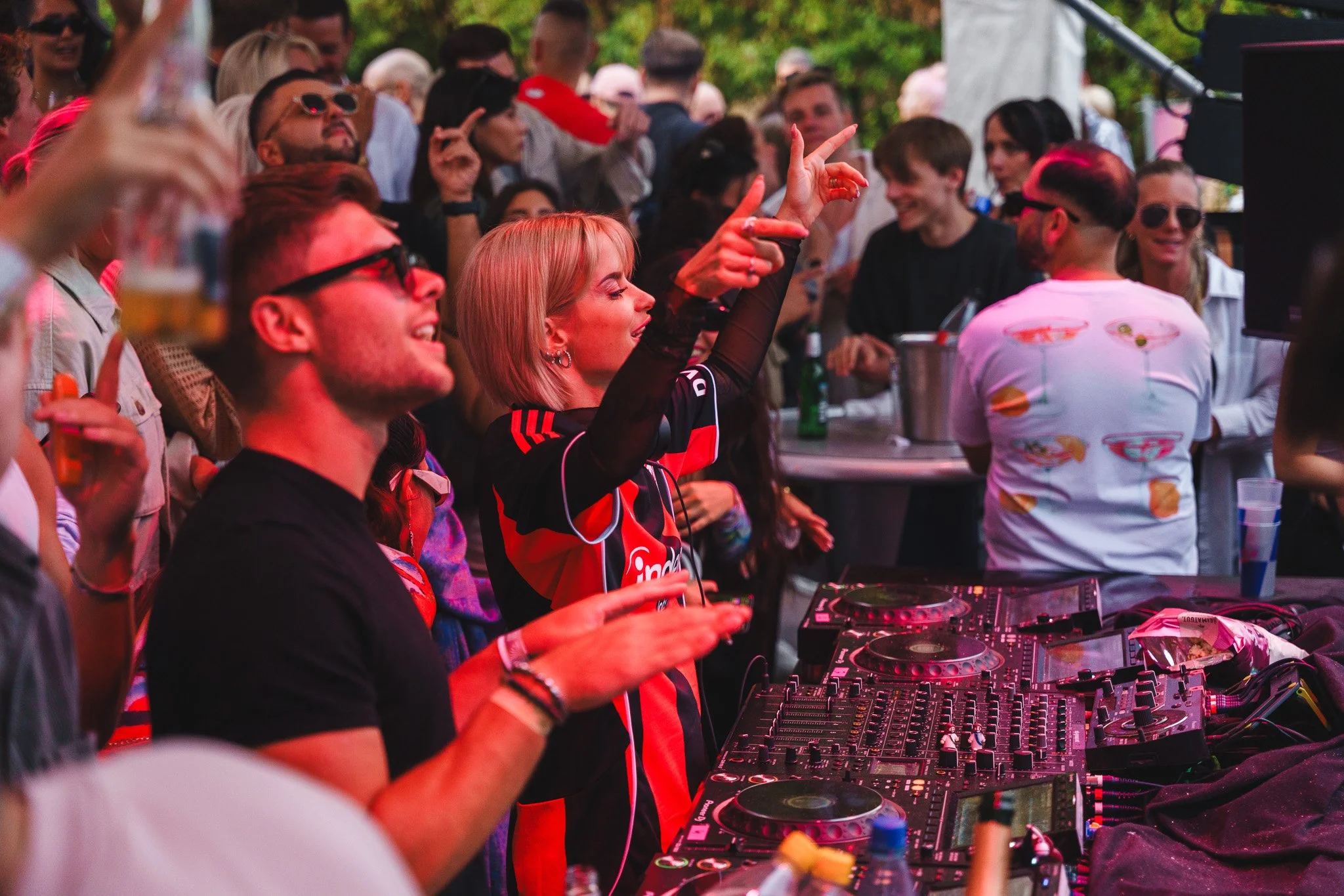 People enjoying music at an outdoor party with a DJ in the foreground and a crowd dancing behind.