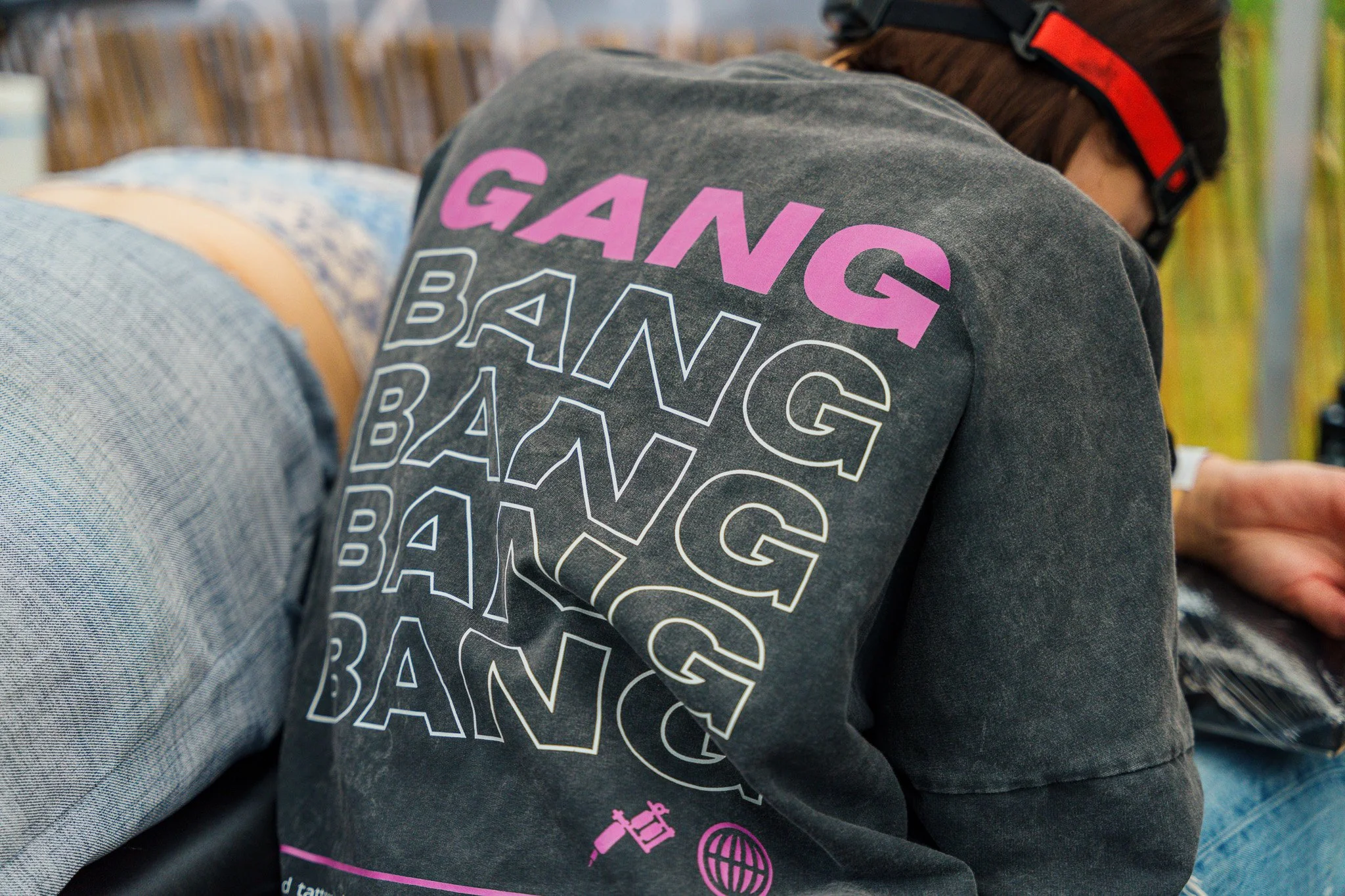 Person wearing a black T-shirt with the word 'GANG' in purple and the word 'BANG' repeated multiple times in white. The person is sitting, leaning over, with their head turned to the side, wearing a black cap with a red stripe. The background shows w