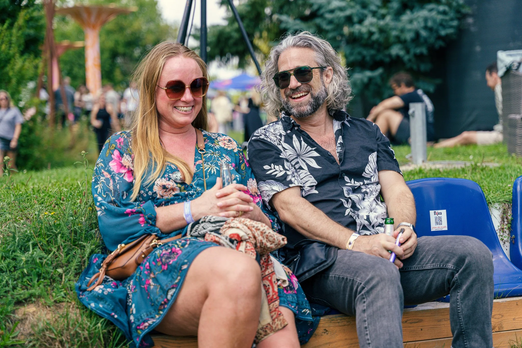 A smiling woman with long red hair and sunglasses sitting outdoors on a bench next to a man with curly gray hair and sunglasses at a festival, both holding drinks.