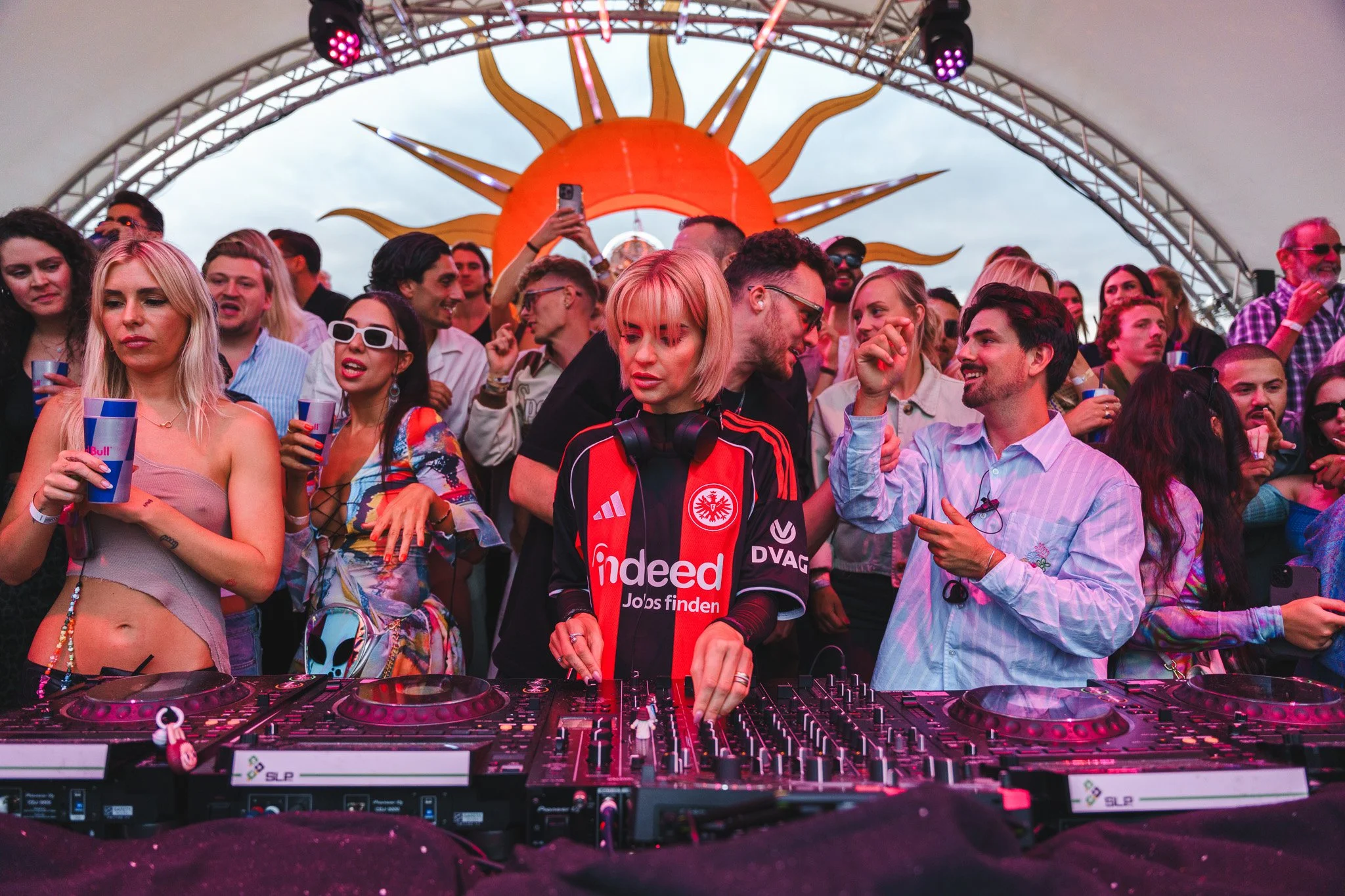 A DJ with short blonde hair wearing an Eintracht Frankfurt jacket is performing at an outdoor music festival with a crowd of people dancing and enjoying the event. The DJ is using a mixing console, and a large sun-shaped decoration is visible in the 