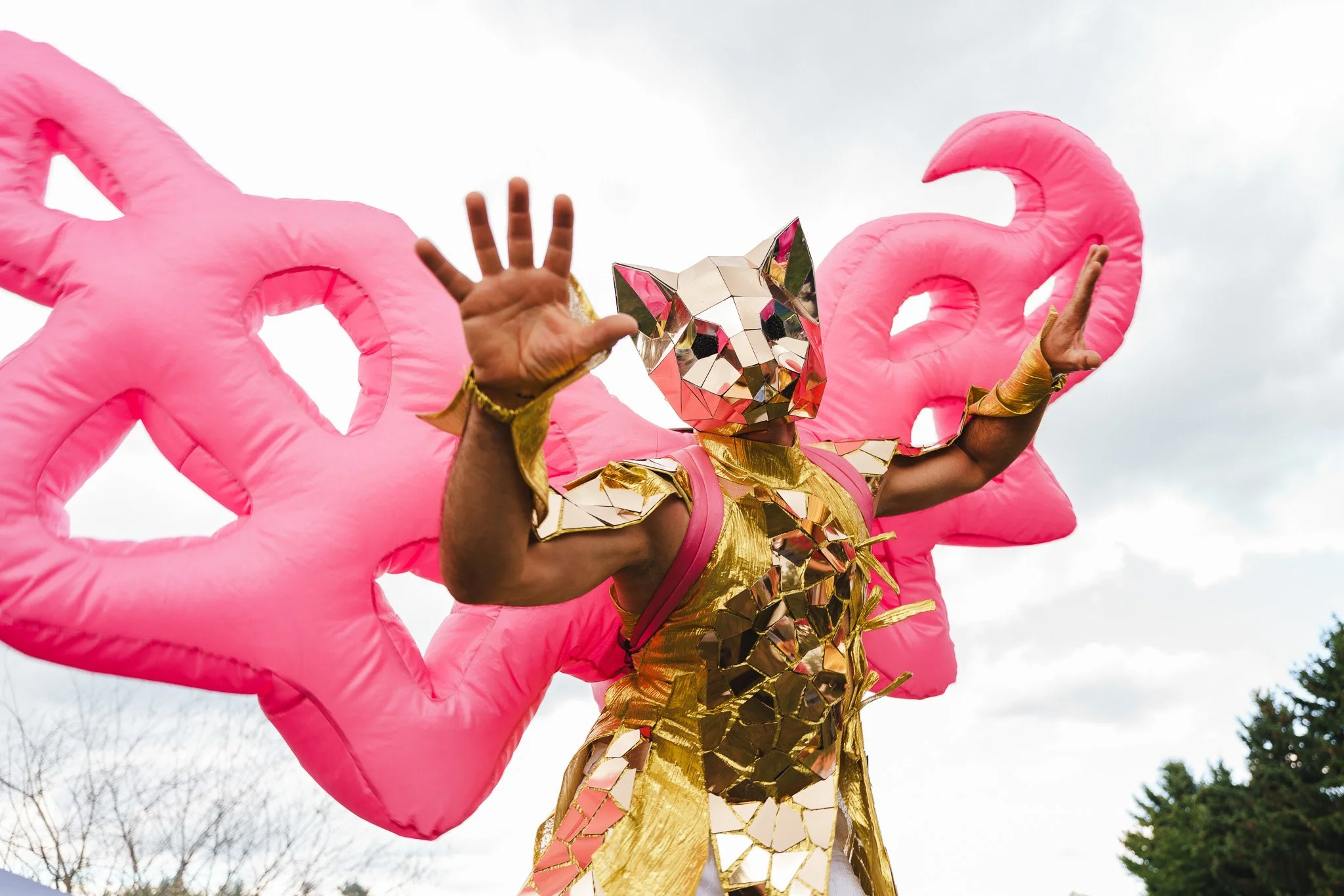 Person wearing a gold and mirror-faceted costume with a pink backpack, posing in front of large pink heart-shaped balloons