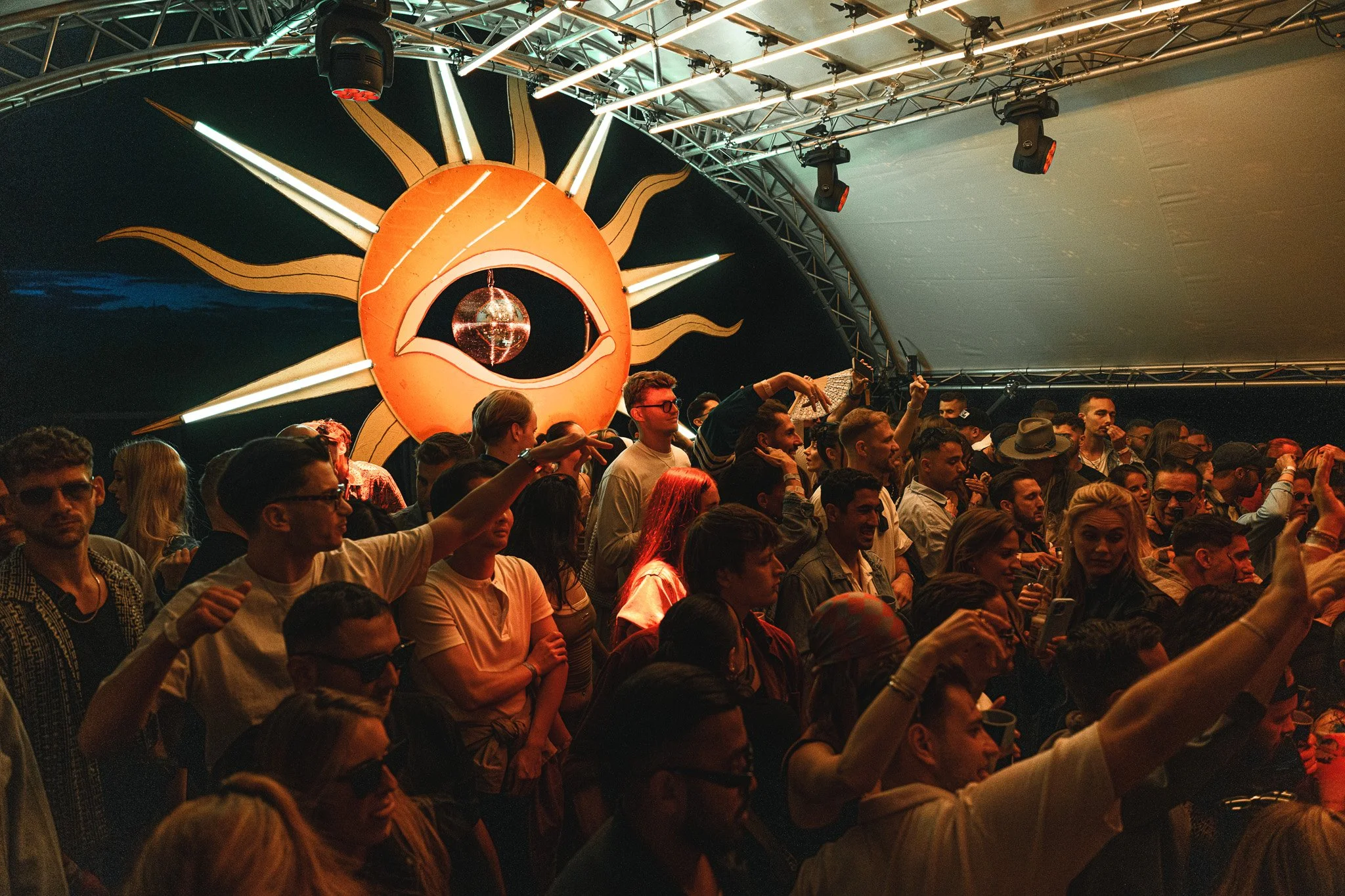 A crowded indoor music event with a large, artistic sun and eye mural on the back wall, people dancing and enjoying the concert, some wearing sunglasses and hats.