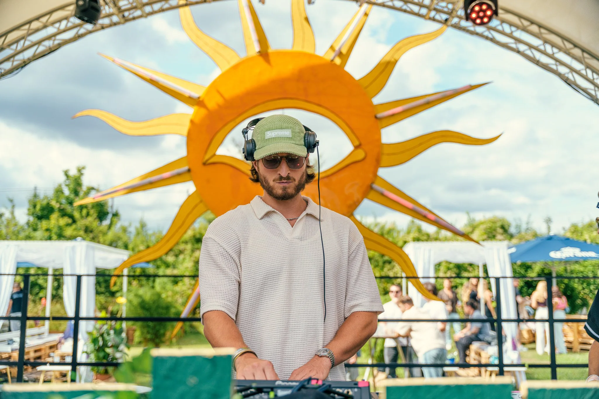 A man DJing at an outdoor festival, with a sun-shaped decoration behind him, wearing sunglasses, headphones, and a green cap.