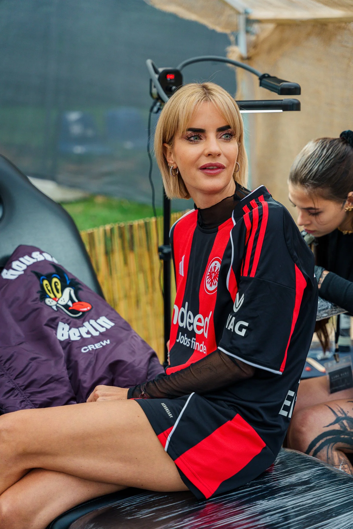 A woman with blonde hair wearing a black and red sports jersey sitting on a bench, with another woman in the background working on a tattoo.