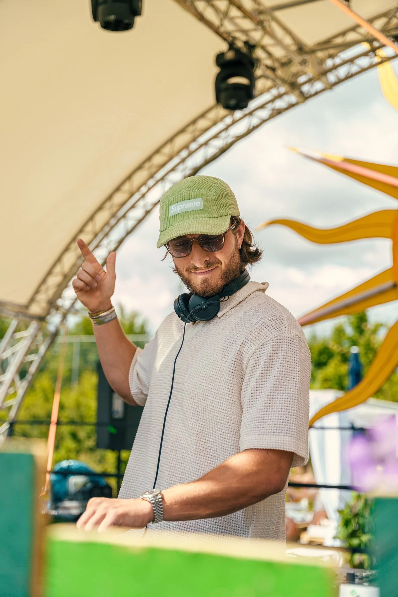 A man wearing a Supreme cap, sunglasses, headphones around his neck, and a watch, smiling and pointing upwards at an outdoor event with a yellow slide and green trees in the background.