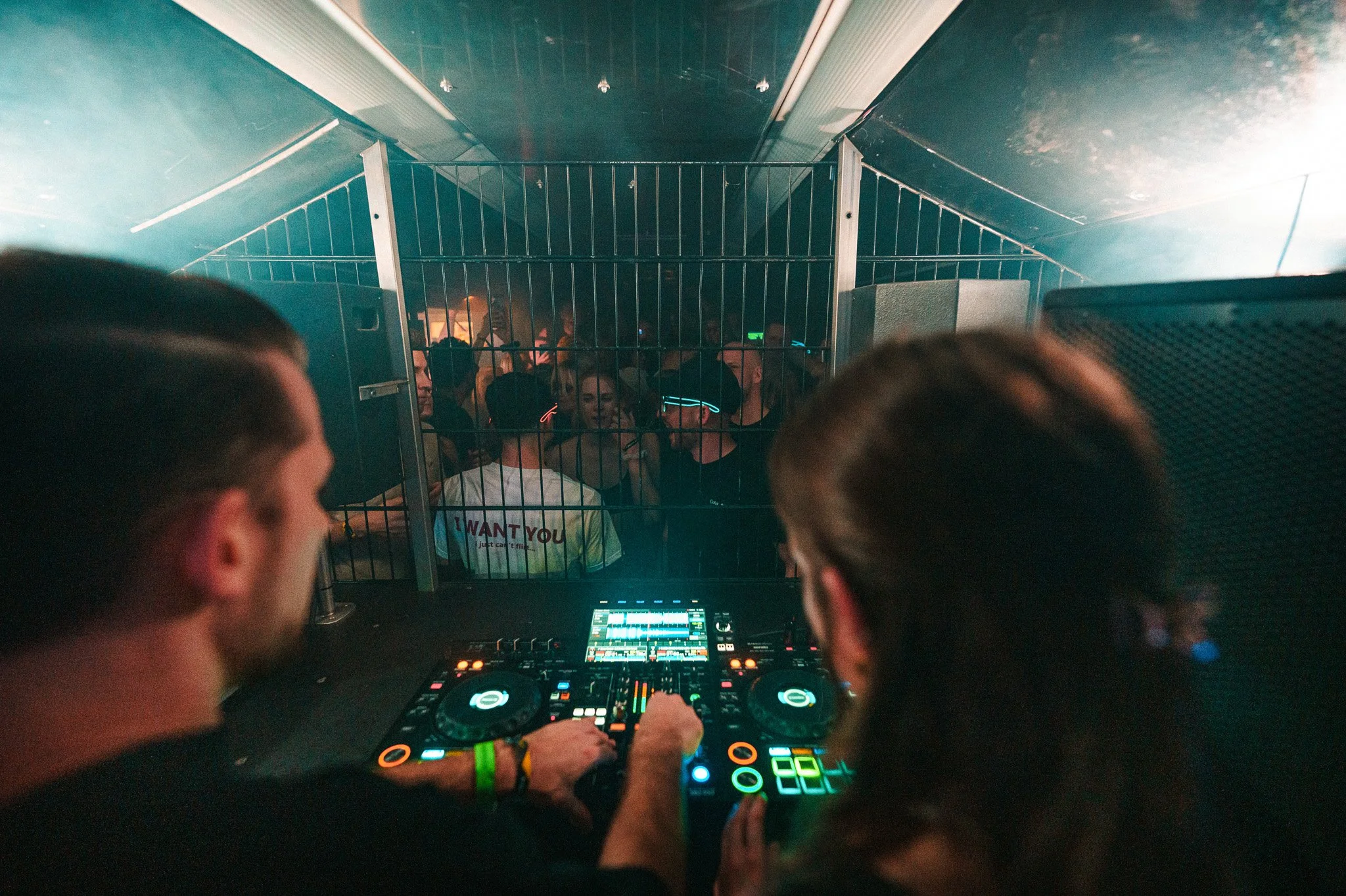 Two DJs are performing at a nightclub, with a crowd dancing behind a metal barrier.