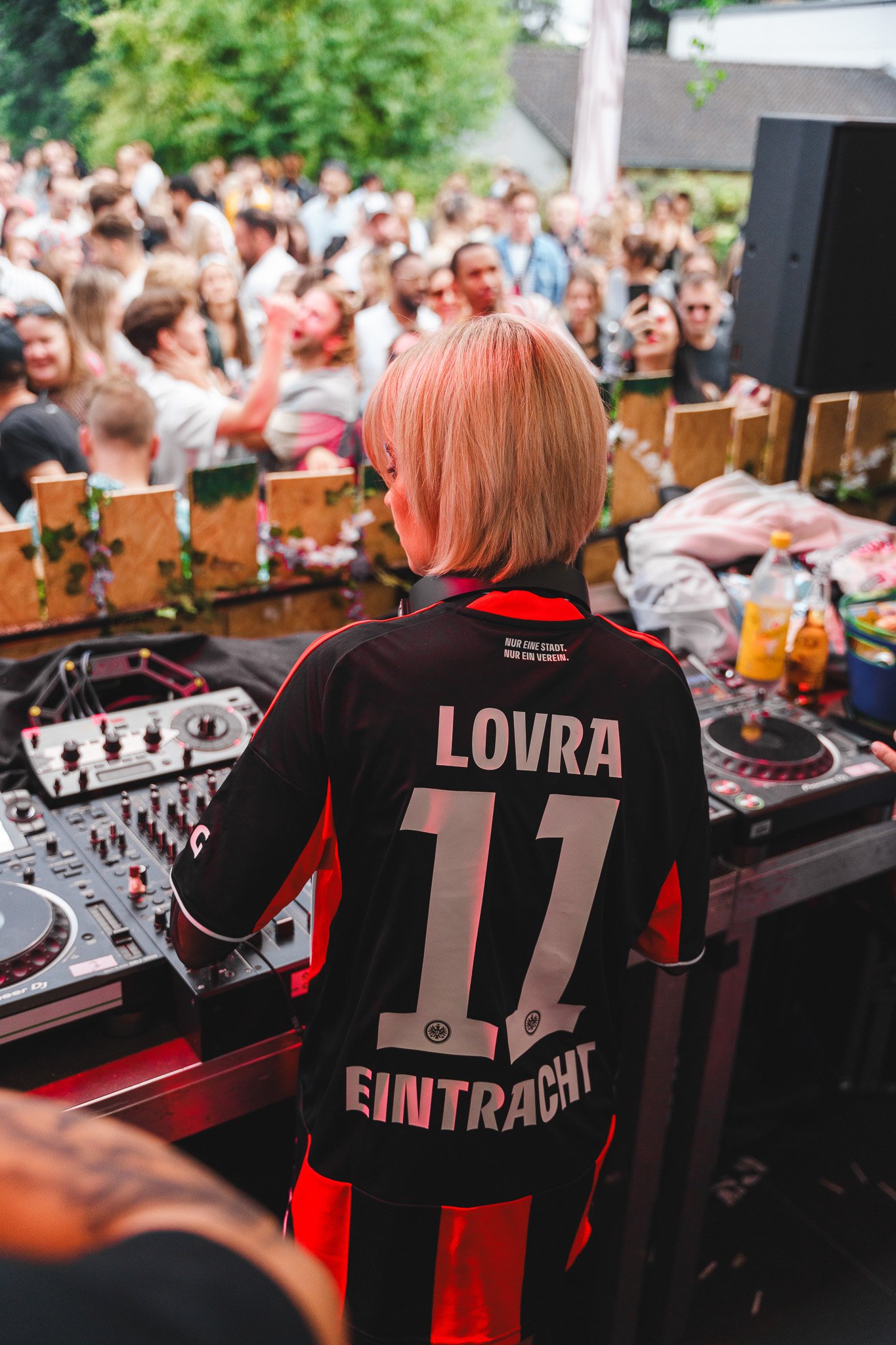 DJ with blonde hair wearing a black and red Eintracht Frankfurt soccer jersey, DJing at an outdoor party with a large crowd of people dancing in the background.