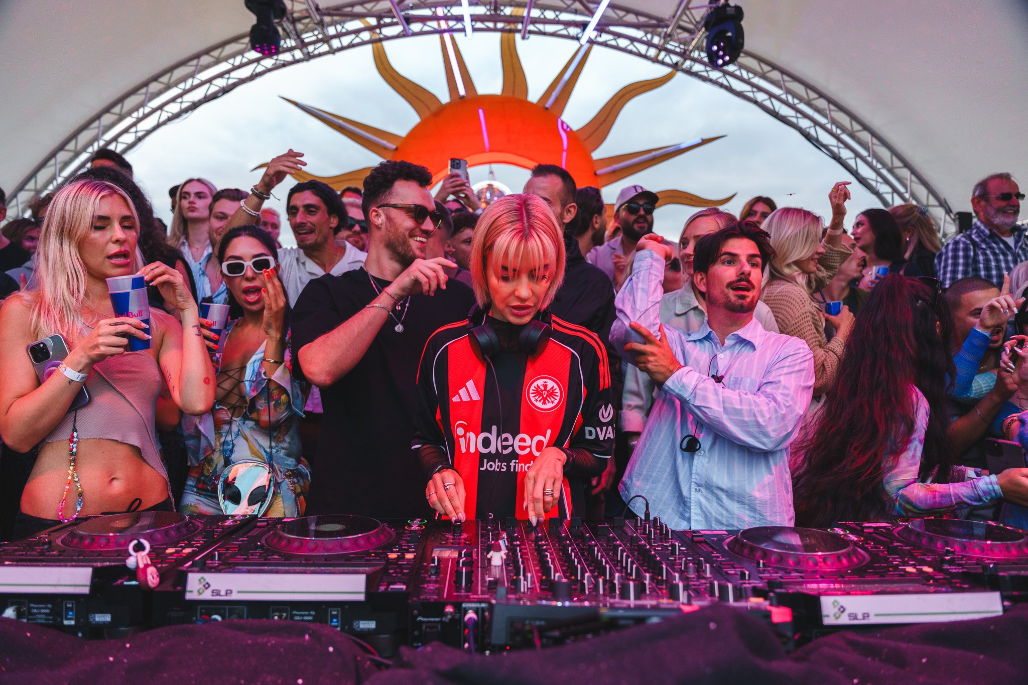 Crowd of people at a music festival or concert, with a DJ woman with pink hair in the center, DJ equipment in front, a large sun-shaped decoration in the background, and many attendees dancing and enjoying the event.
