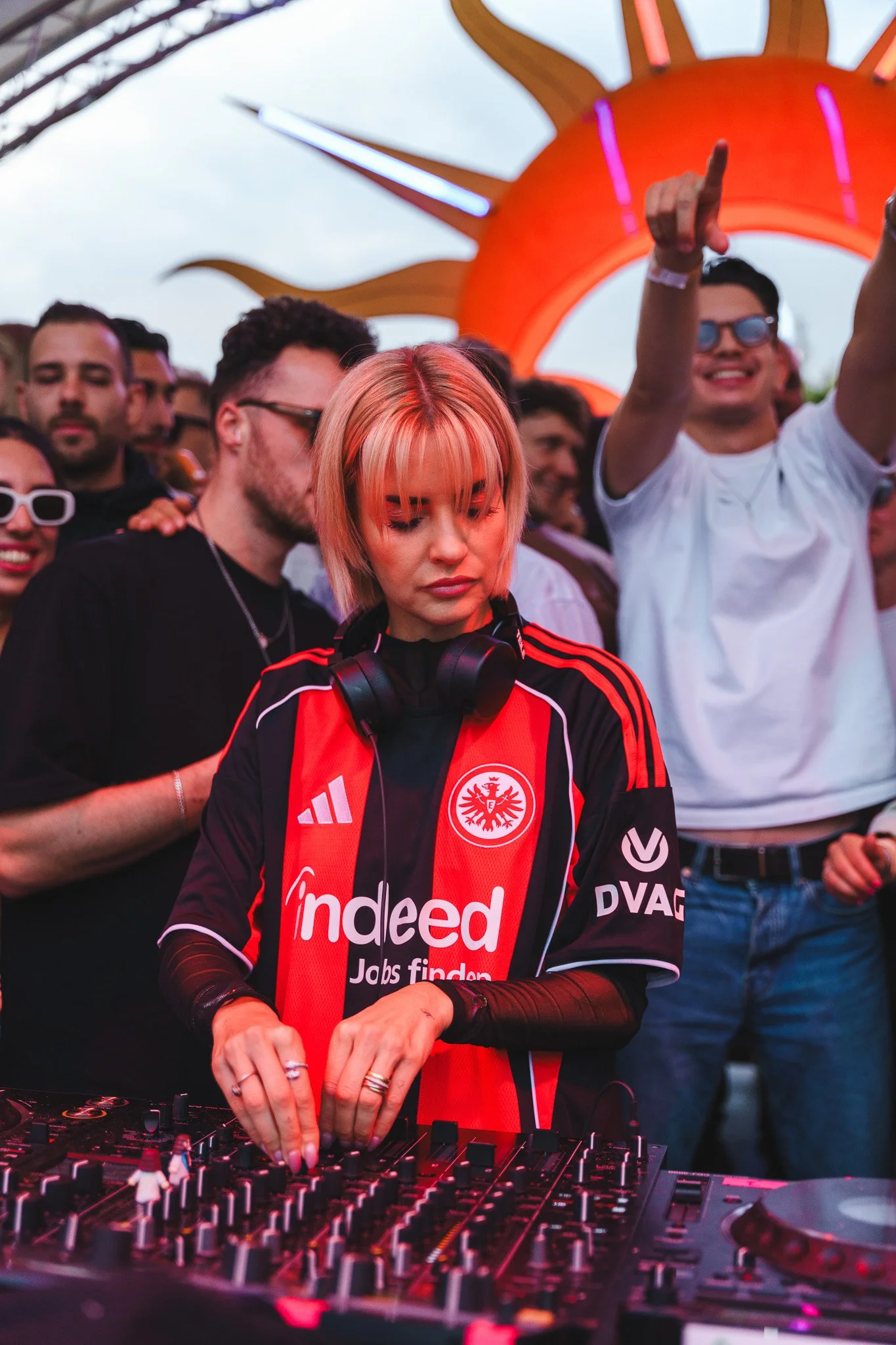 A female DJ with pink hair and headphones around her neck, wearing a red and black soccer jersey with logos, mixes music at an outdoor event with a crowd of people dancing and enjoying the music, and a large orange sun-shaped decoration in the backgr