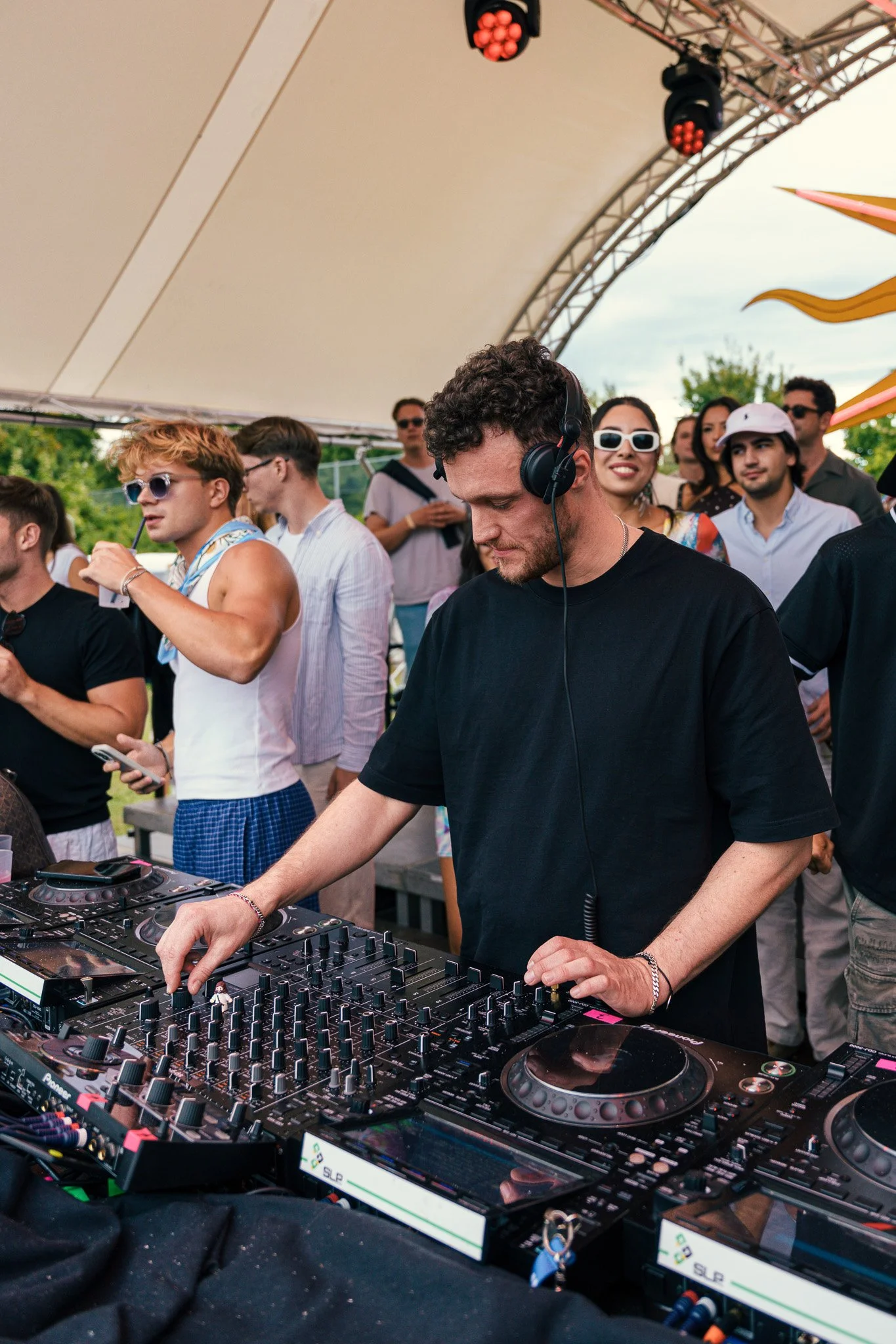A DJ wearing black t-shirt, headphones, and bracelet, is DJing with a mixer and turntables at an outdoor event, with a group of people dancing and enjoying in the background under a large canopy.