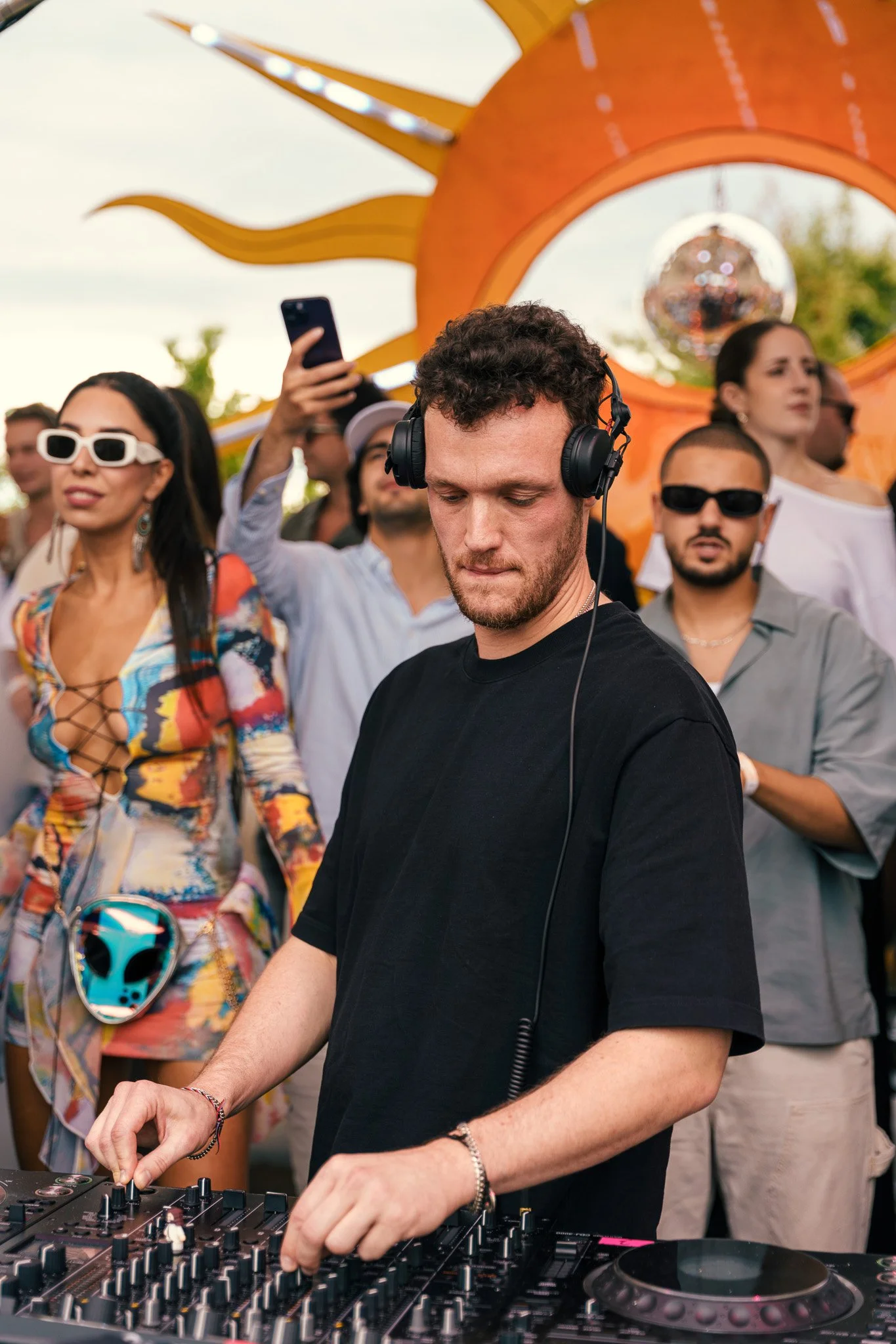A DJ plays music at an outdoor event with a crowd of people dancing and enjoying the atmosphere, featuring a large orange sun-shaped decoration in the background.