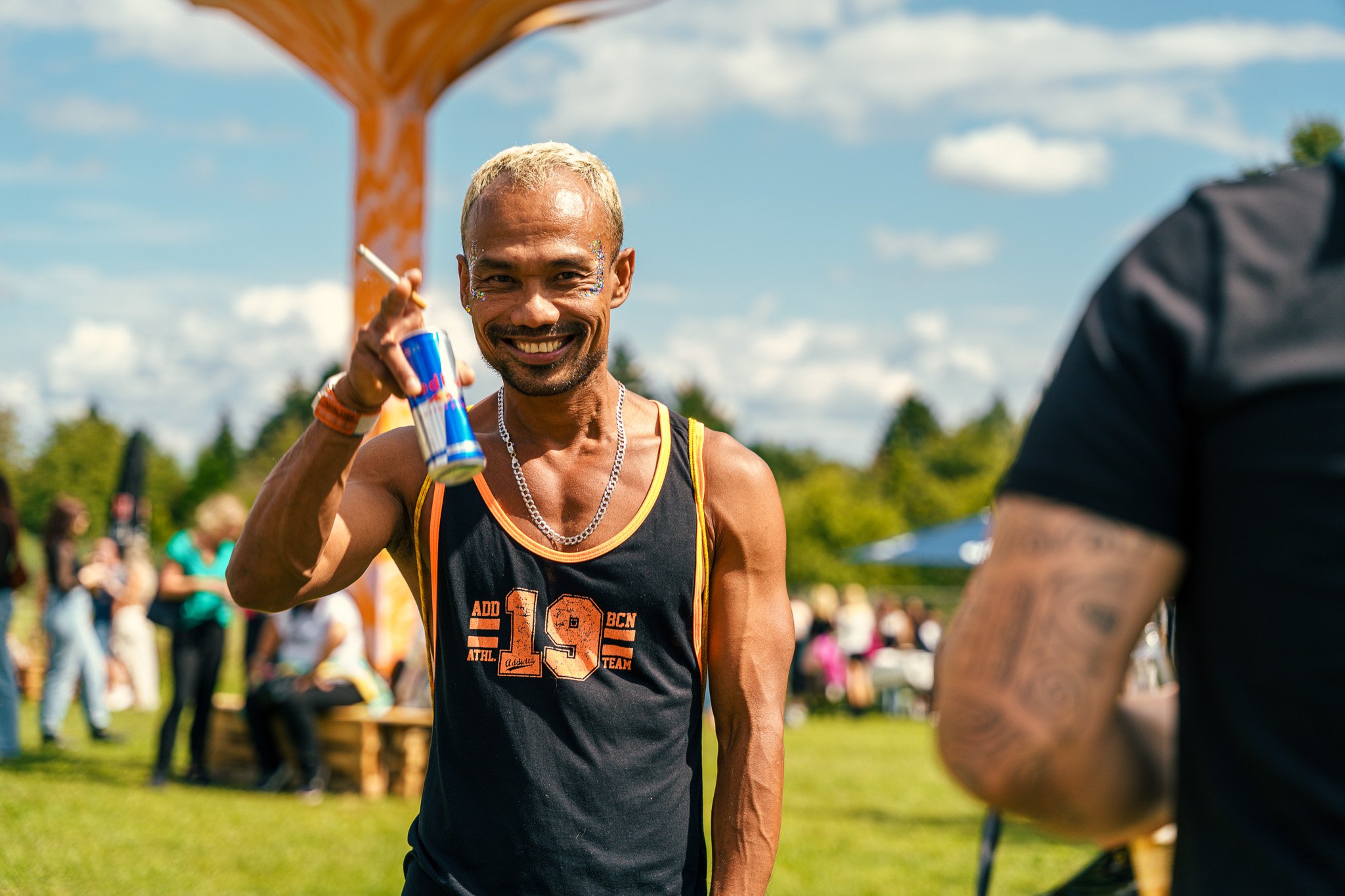 Smiling man with short blond hair and glitter on his face holding a Red Bull can and a cigarette outdoors during a sunny day, with people gathered in the background at a park event.
