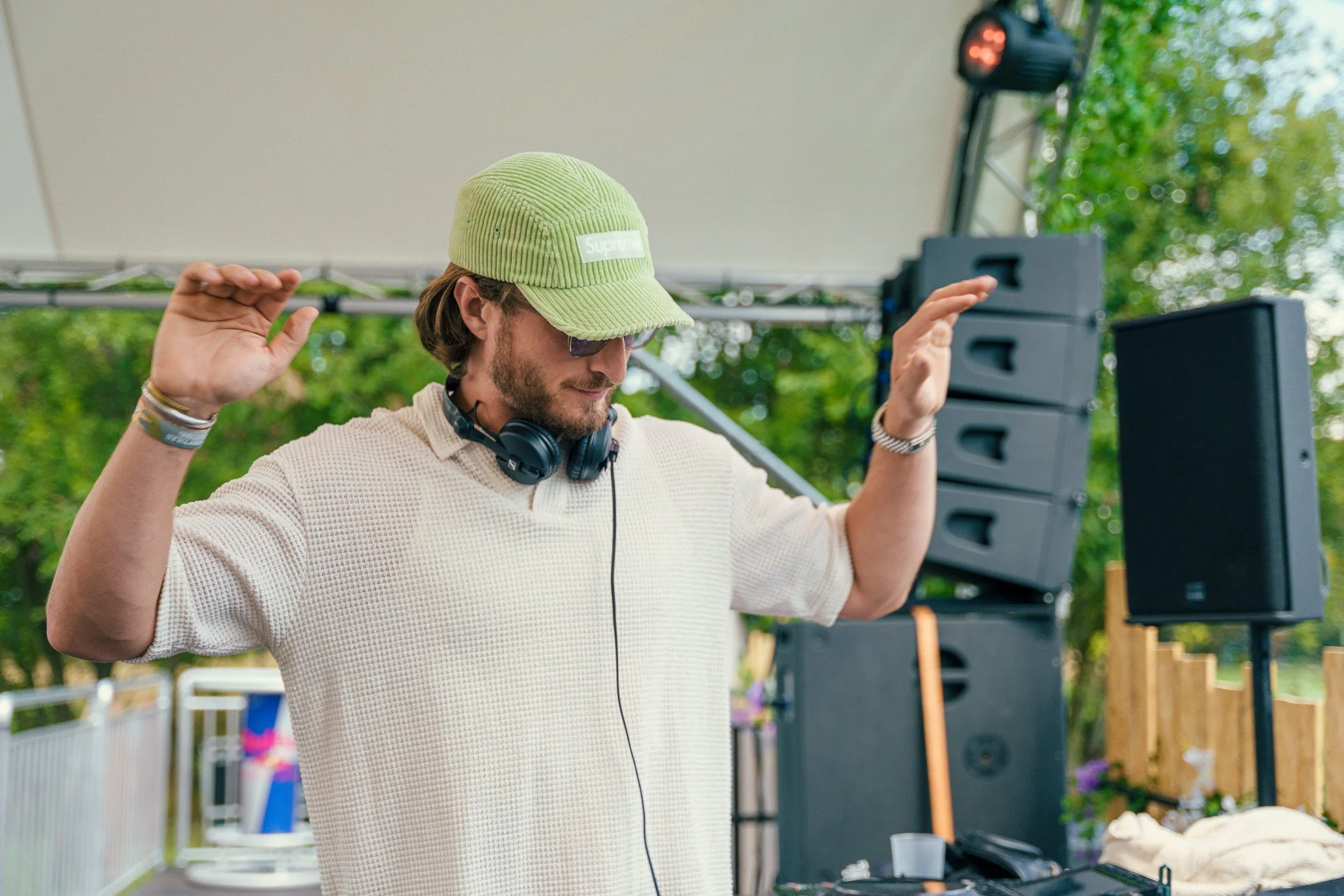 A man DJing outdoors at a festival, wearing a green Supreme cap and sunglasses, with headphones around his neck, raising his hands.