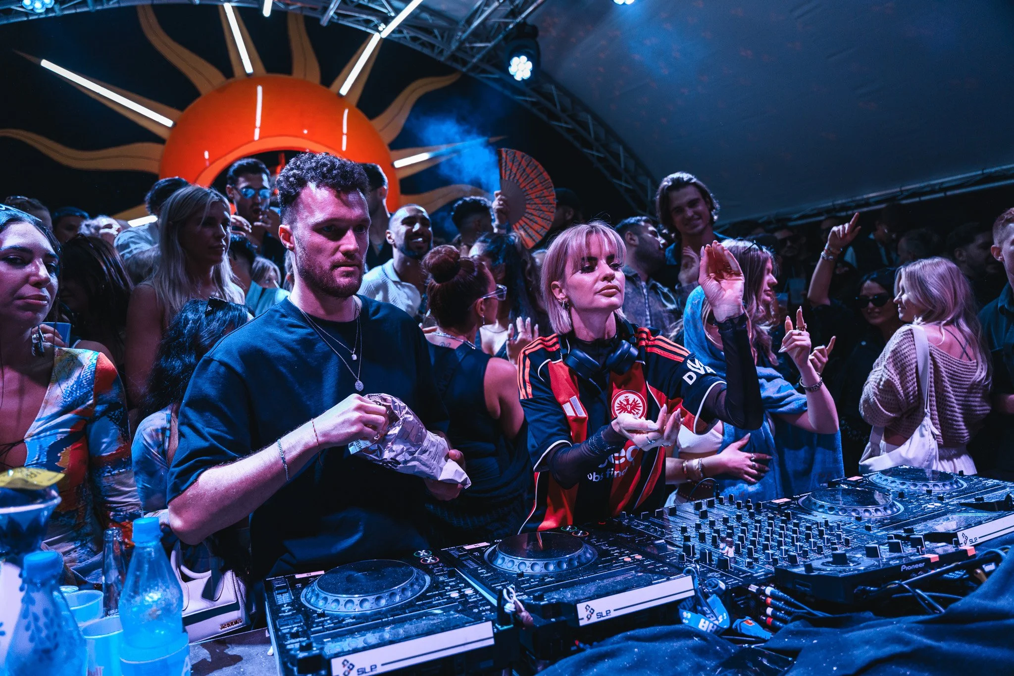 DJ and crowd at a nightclub with a sun and rays decoration overhead.