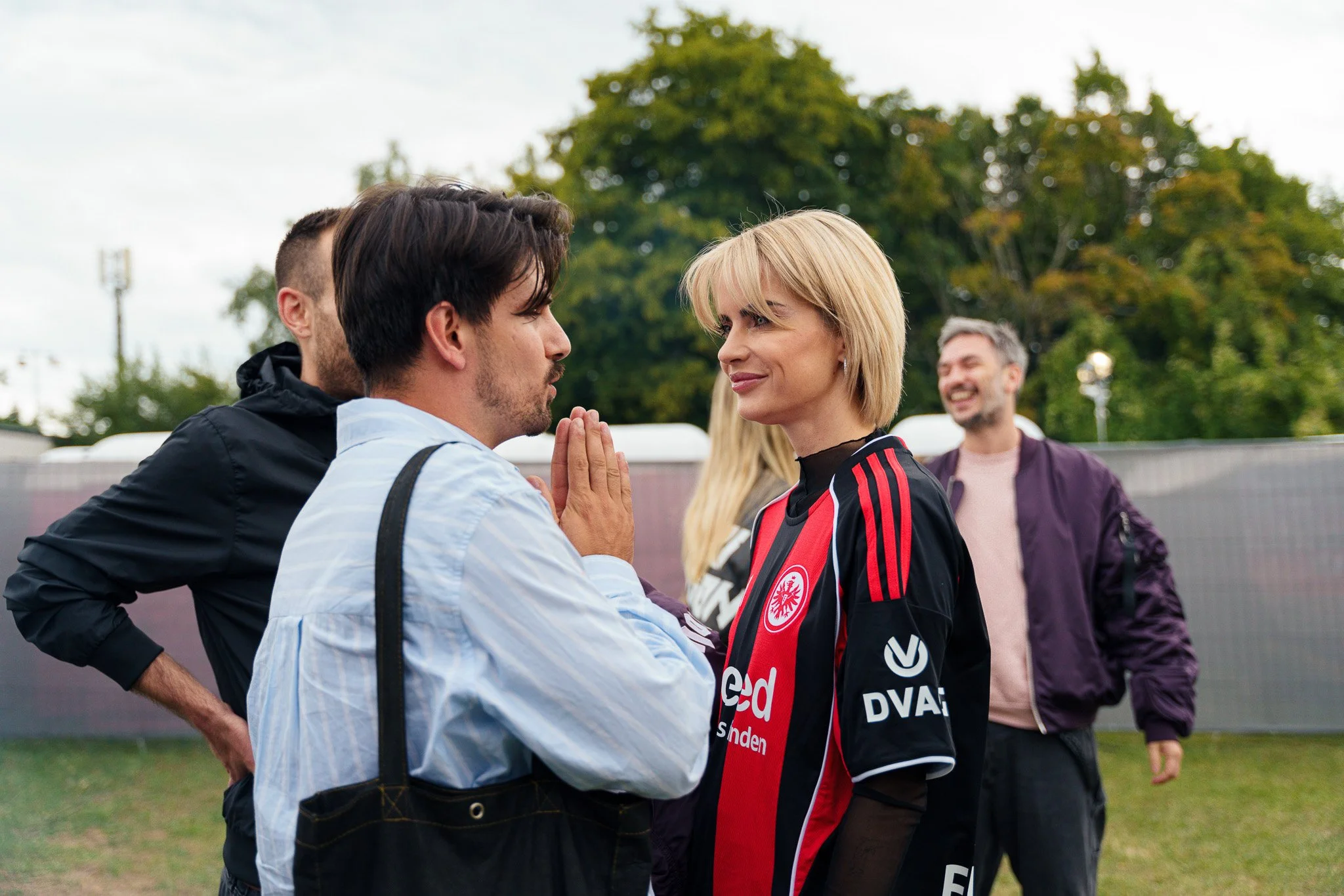 A man and woman face-to-face with hands pressed together, outdoors on a cloudy day, with a man smiling in the background