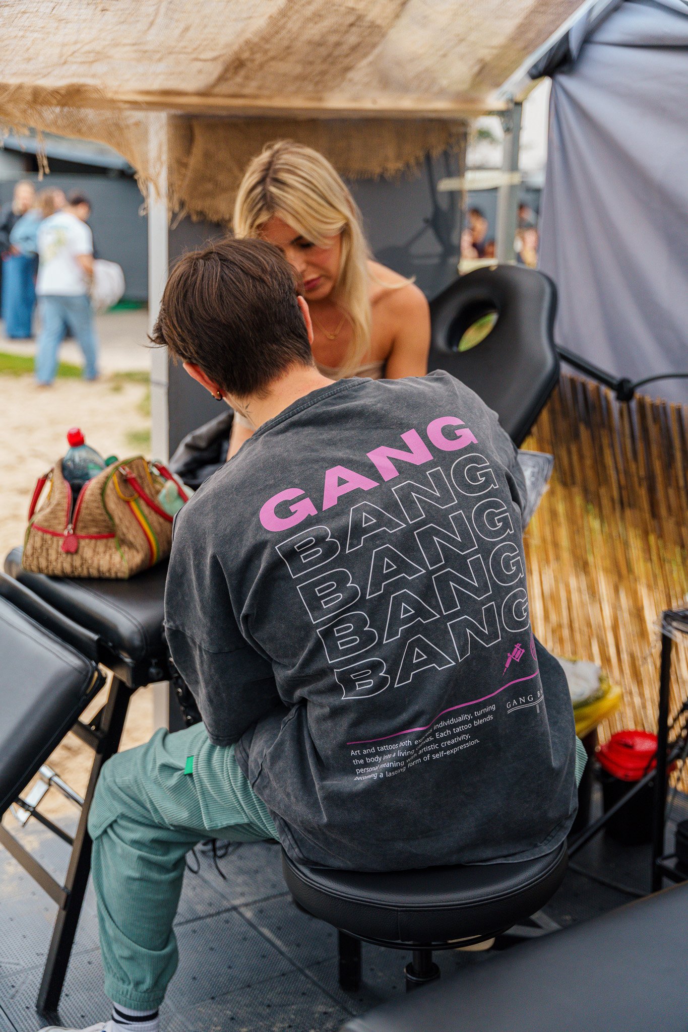 Two women at a tattoo booth, one tattoo artist working on a client. The tattoo artist is wearing a black T-shirt with the word 'GANG' in purple and repeated multiple times on the back. The client has long blonde hair and is seated with her back expos