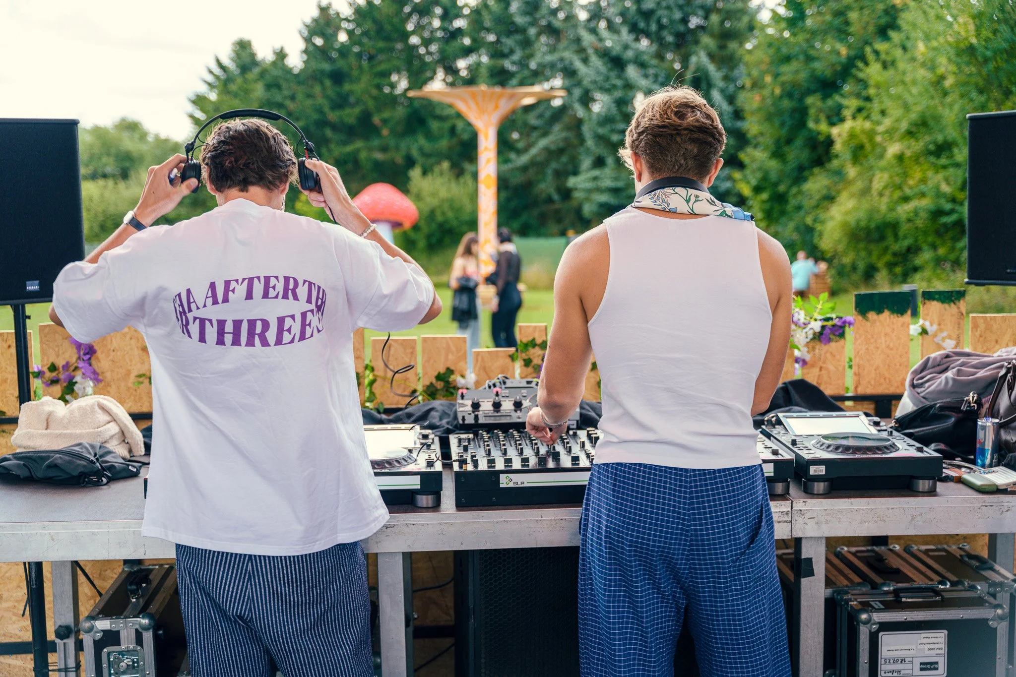 Two men DJing outdoors with a colorful attraction in the background and a lush green setting.