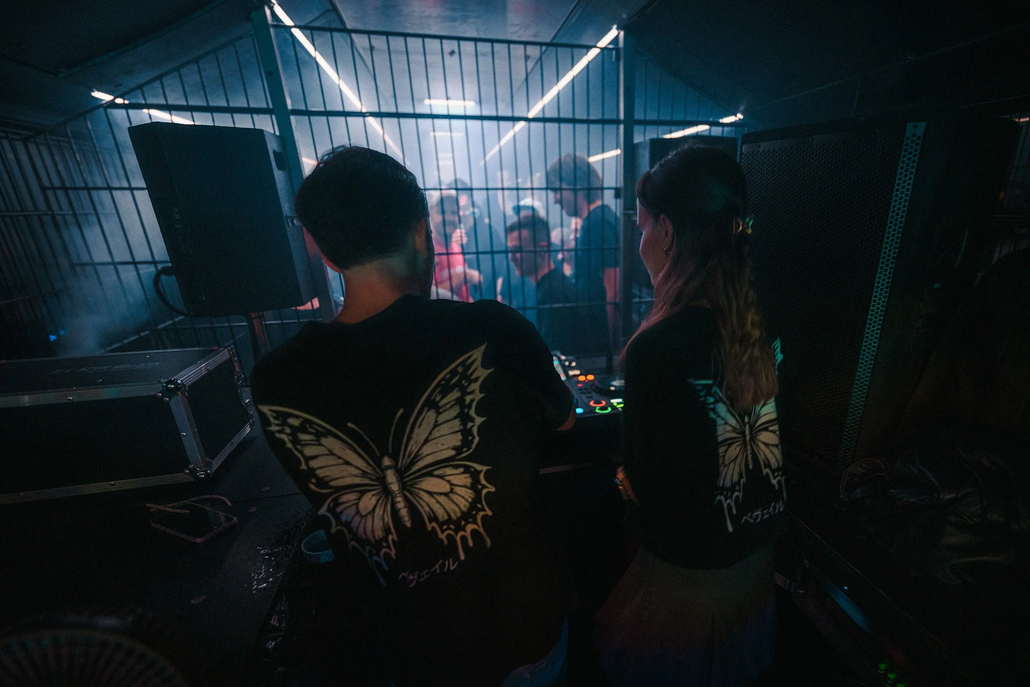 Two DJs are performing at a crowded nightclub with people dancing behind a metal fence.