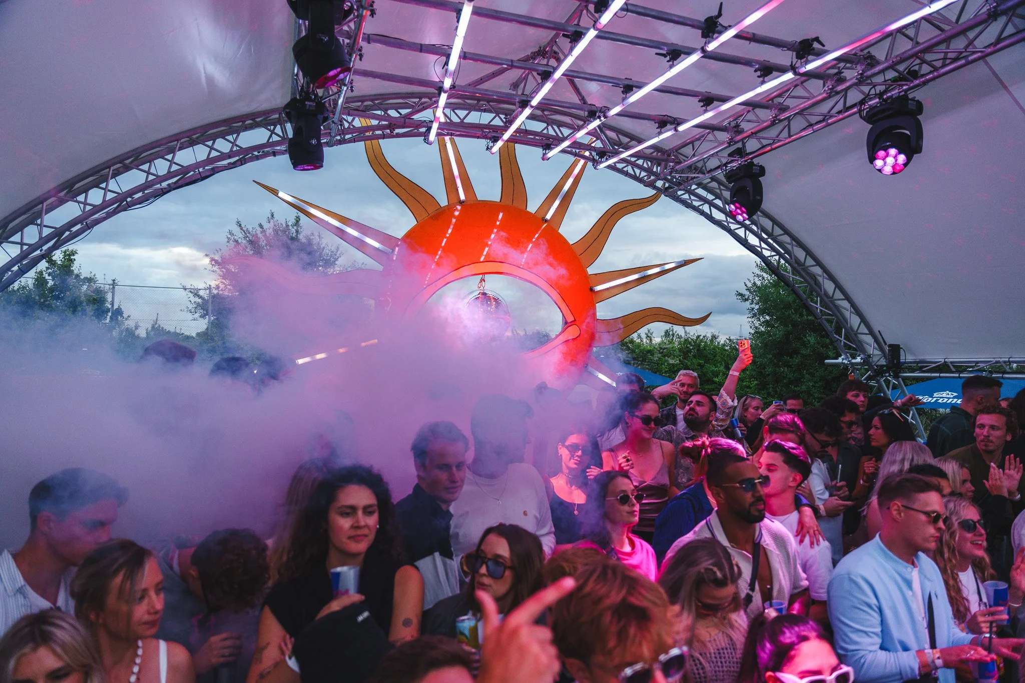 Crowd of people dancing and having fun at an outdoor music festival with a large, decorative sun and fog effects.