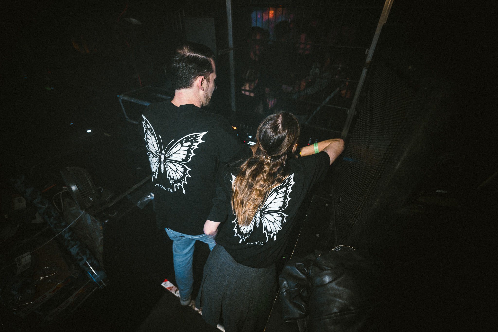 Two DJs with butterfly graphics on their black shirts performing at a nightclub, with a crowd and DJ equipment in the background.