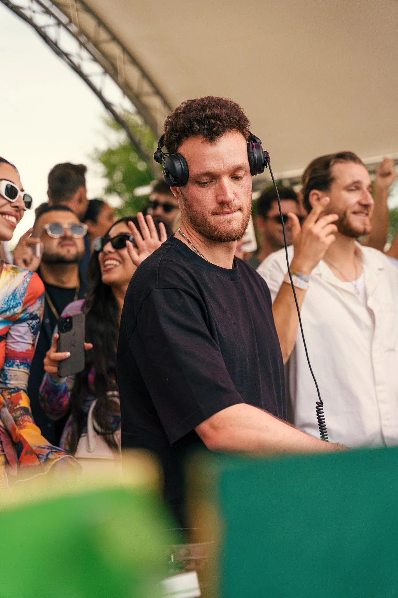 A DJ with headphones on is playing music at an outdoor event, surrounded by a crowd of people enjoying the music and taking photos.