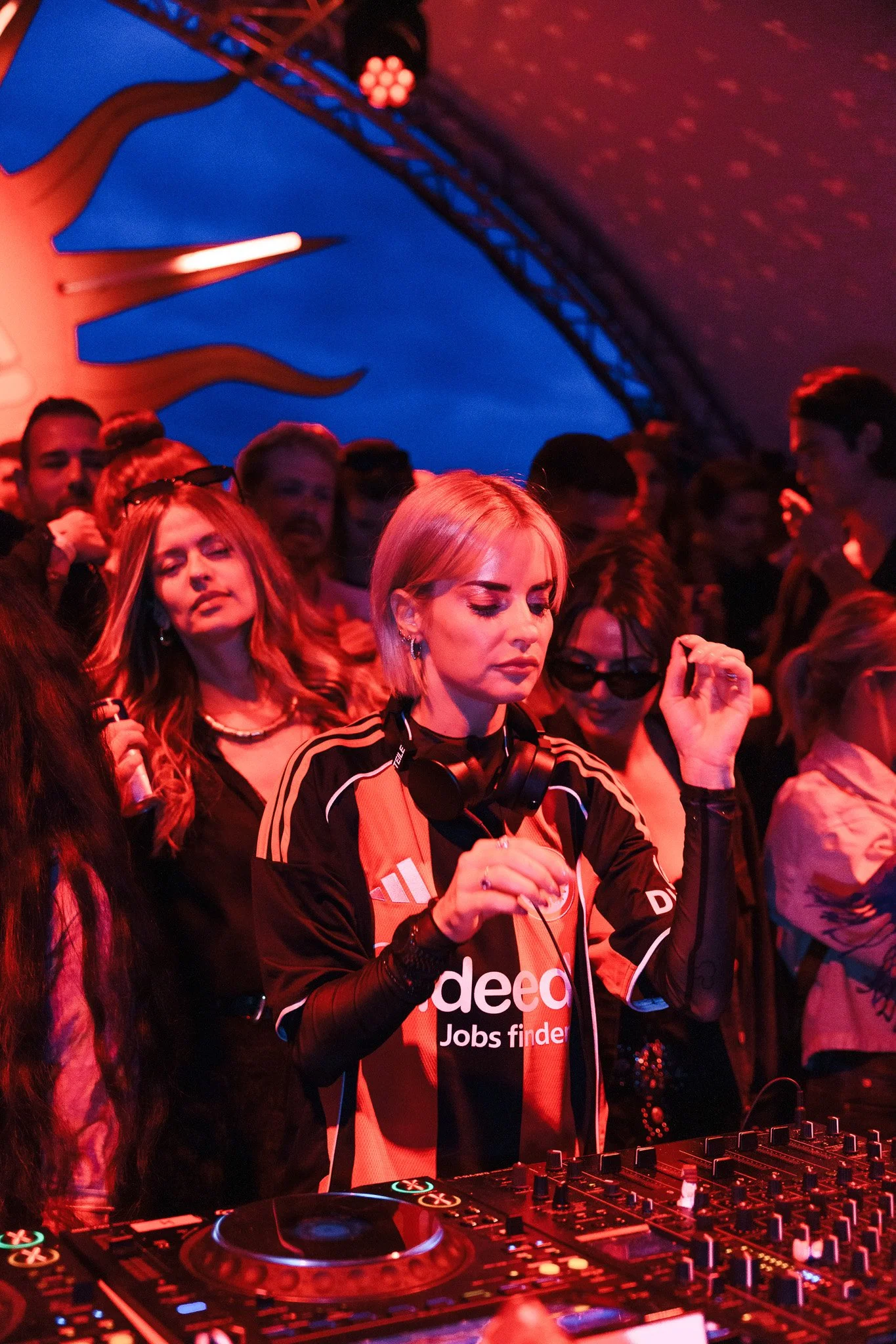 A female DJ wearing a black and red sports jersey, headphones around her neck, is DJing at a crowded event with colorful lighting. She has short blonde hair and is focused on her equipment, with a group of people dancing and enjoying music in the bac