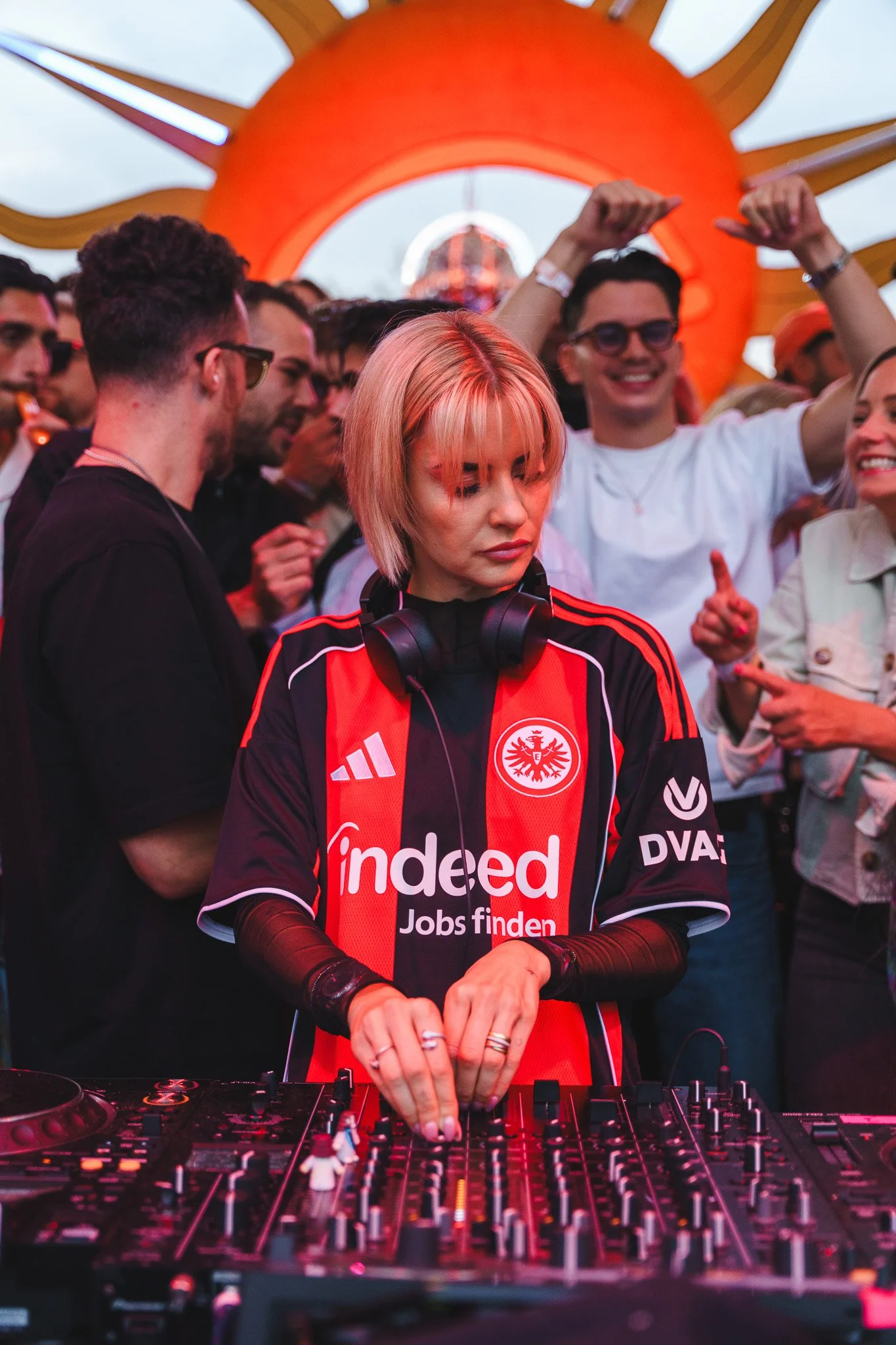 A female DJ with short blonde hair wearing large headphones and a red and black FC Eintracht Frankfurt jersey, DJing at a crowded outdoor event with a large orange sun decoration in the background.