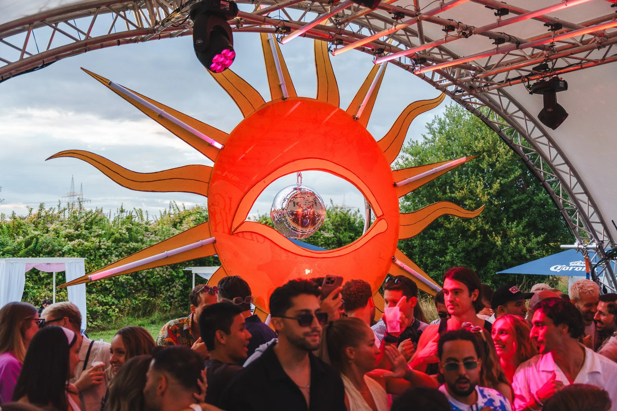 Crowd of people at an outdoor event with a large decorative sun with an eye and a disco ball in the center.
