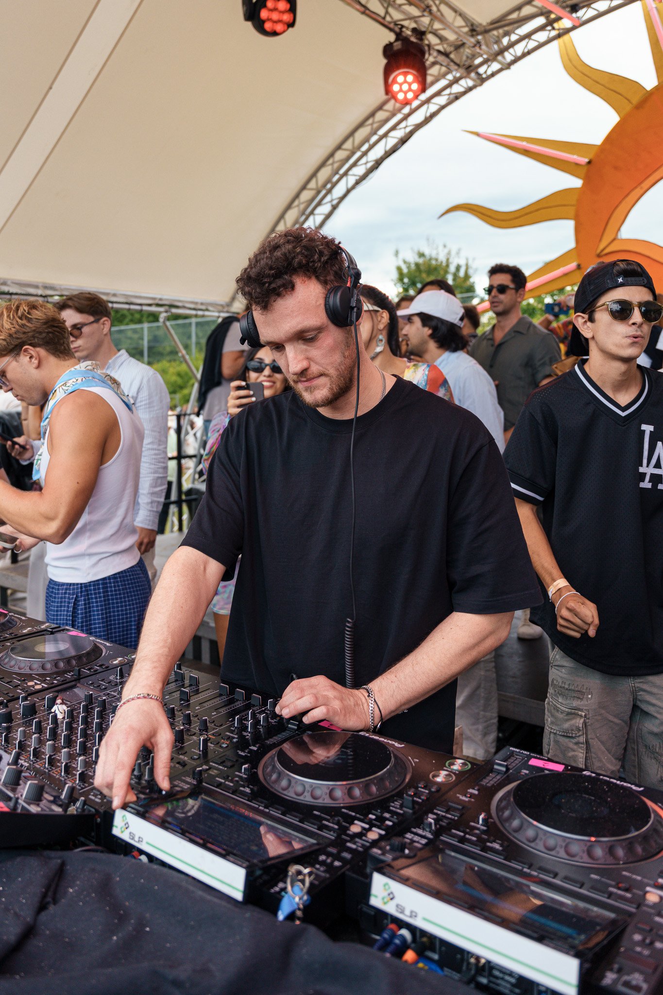 A DJ with curly hair wearing headphones, dressed in a black t-shirt, performing with DJ equipment at an outdoor event. There are people in the background, some taking photos, under a tent with stage lighting and a decorative sun element.