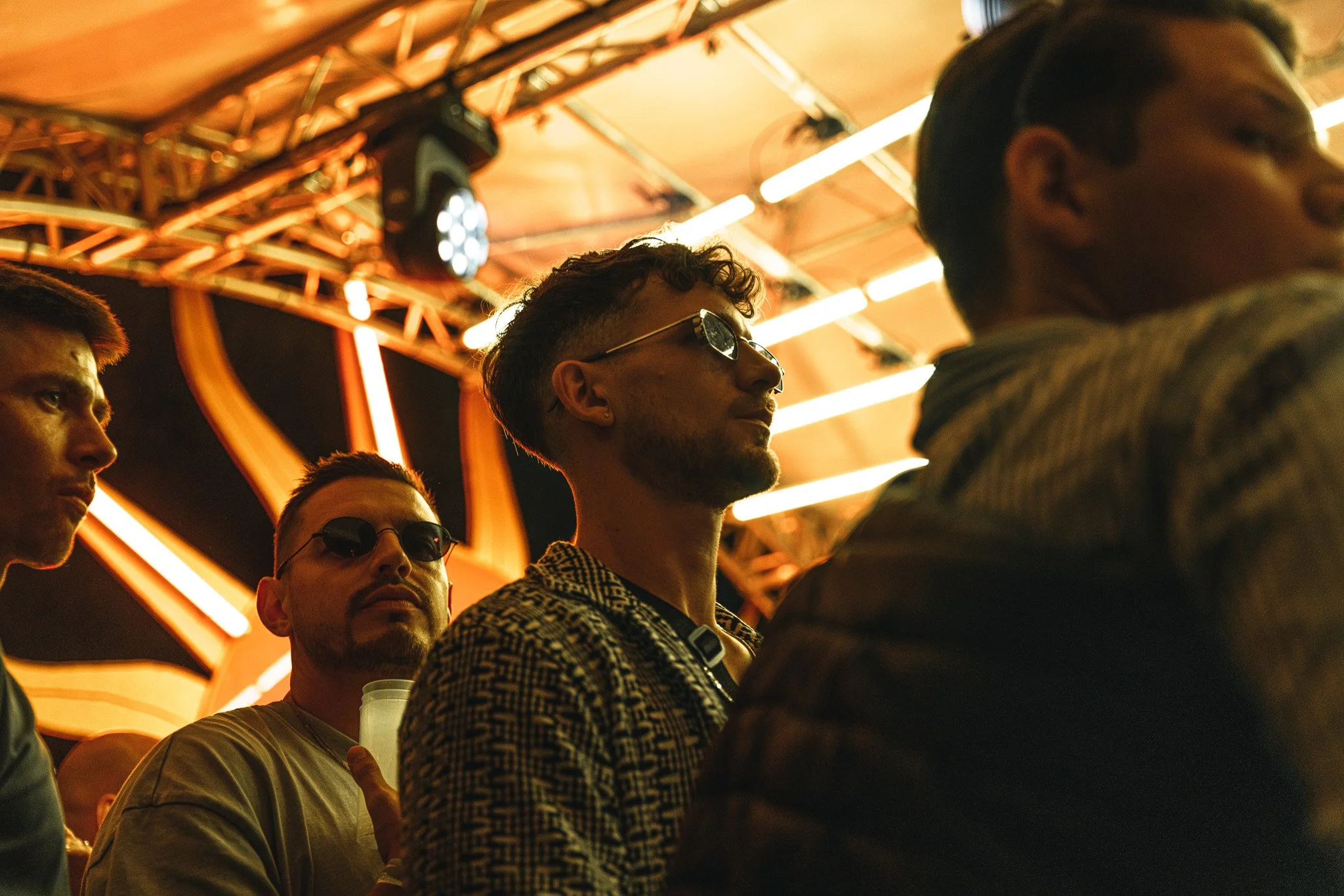A group of young men sitting in a row at a dimly lit venue with orange lighting and neon signs, some wearing sunglasses.
