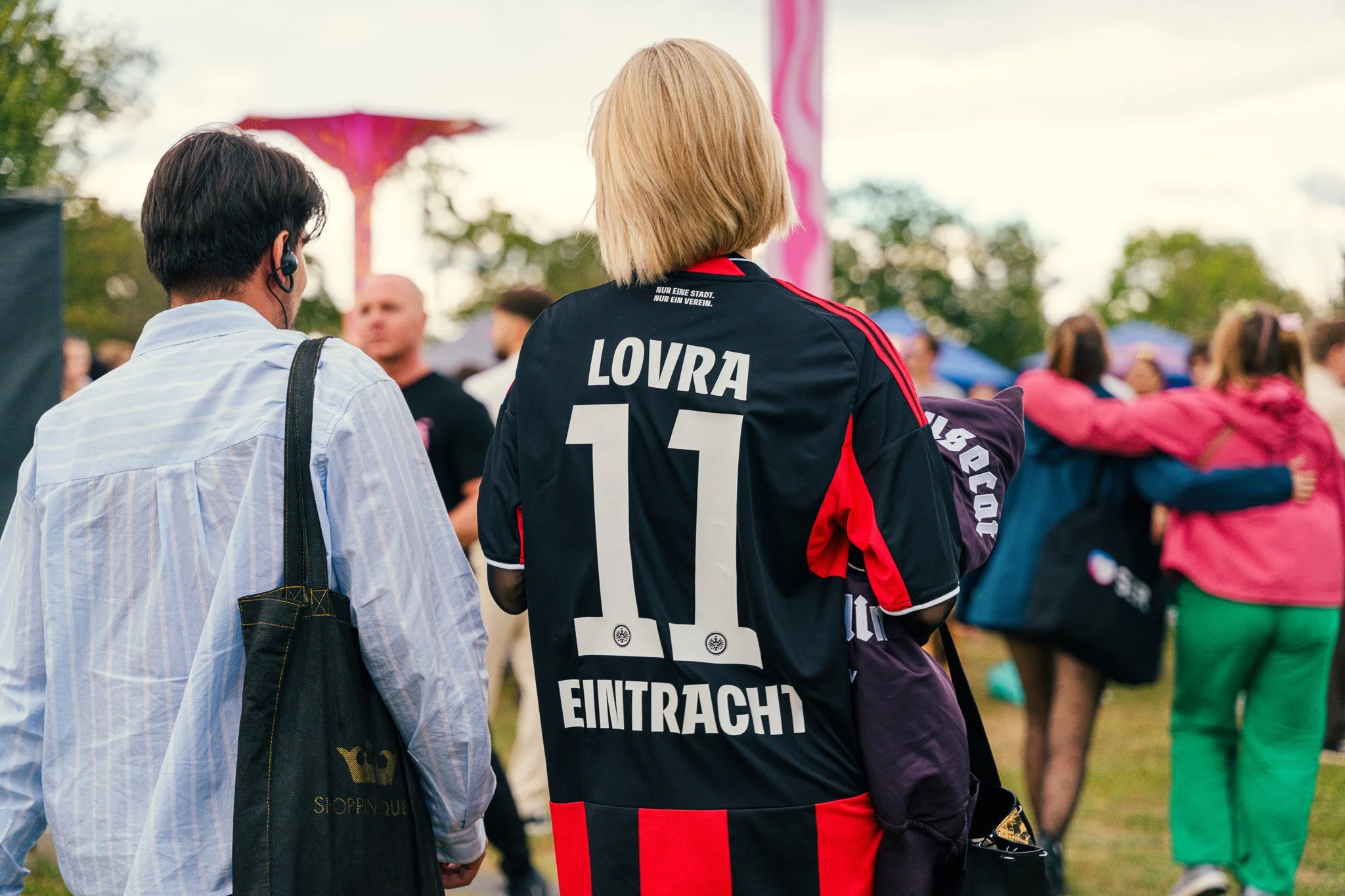 Back view of a blonde woman wearing a black and red soccer jersey with the name 'LOVRA' and the number '11' on it, standing outdoors among a crowd of people.