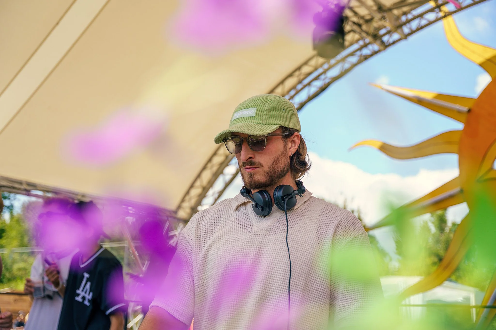 A man with sunglasses, a green cap, and headphones around his neck stands outdoors under a large canopy, with colorful flower decorations nearby.