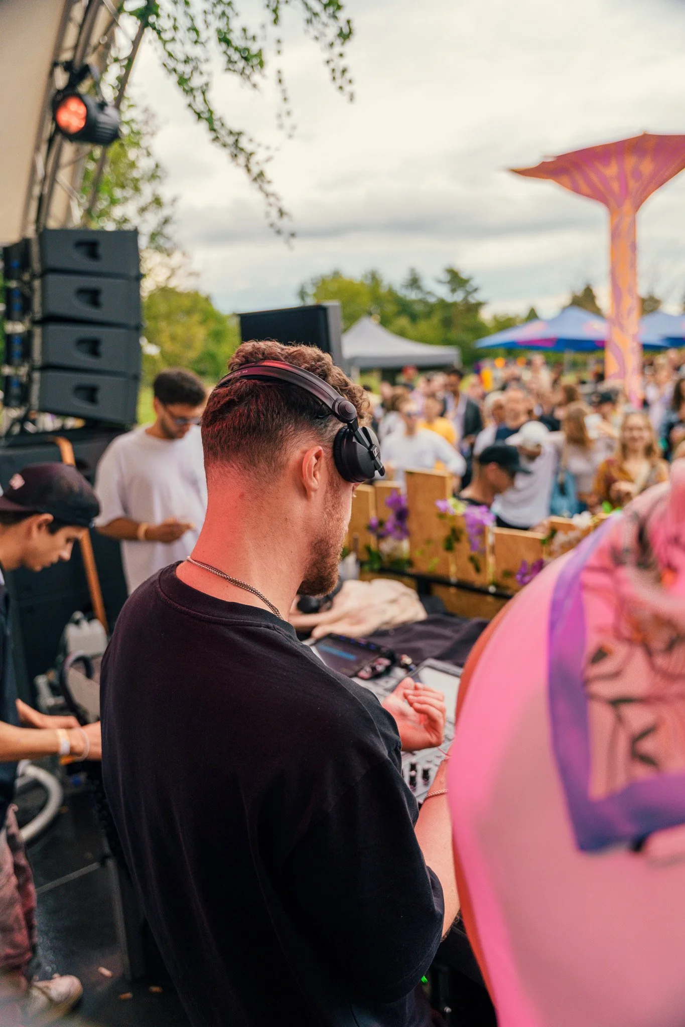 DJ wearing headphones at an outdoor music event, with a crowd of people, tents, and trees in the background.
