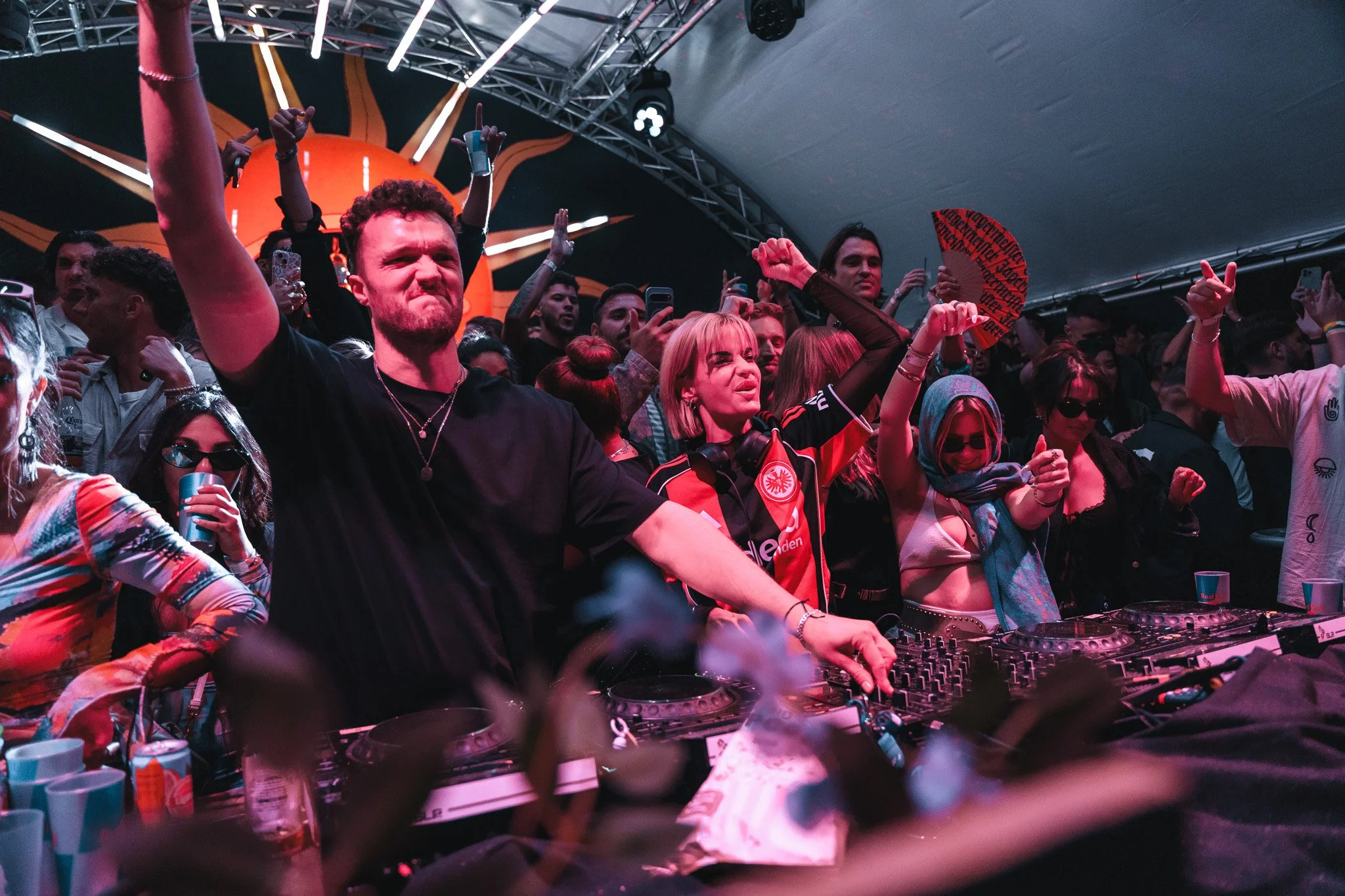 DJ or music producer with blonde hair, wearing headphones around neck, performing at a DJ booth with a crowd of people dancing and raising their hands under a tent with stage lights.