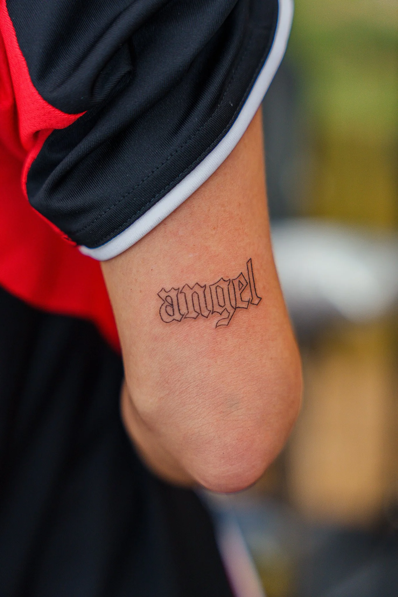 Tattoo of the word 'angel' in Gothic font on a person's arm, with the person's clothing partially visible.