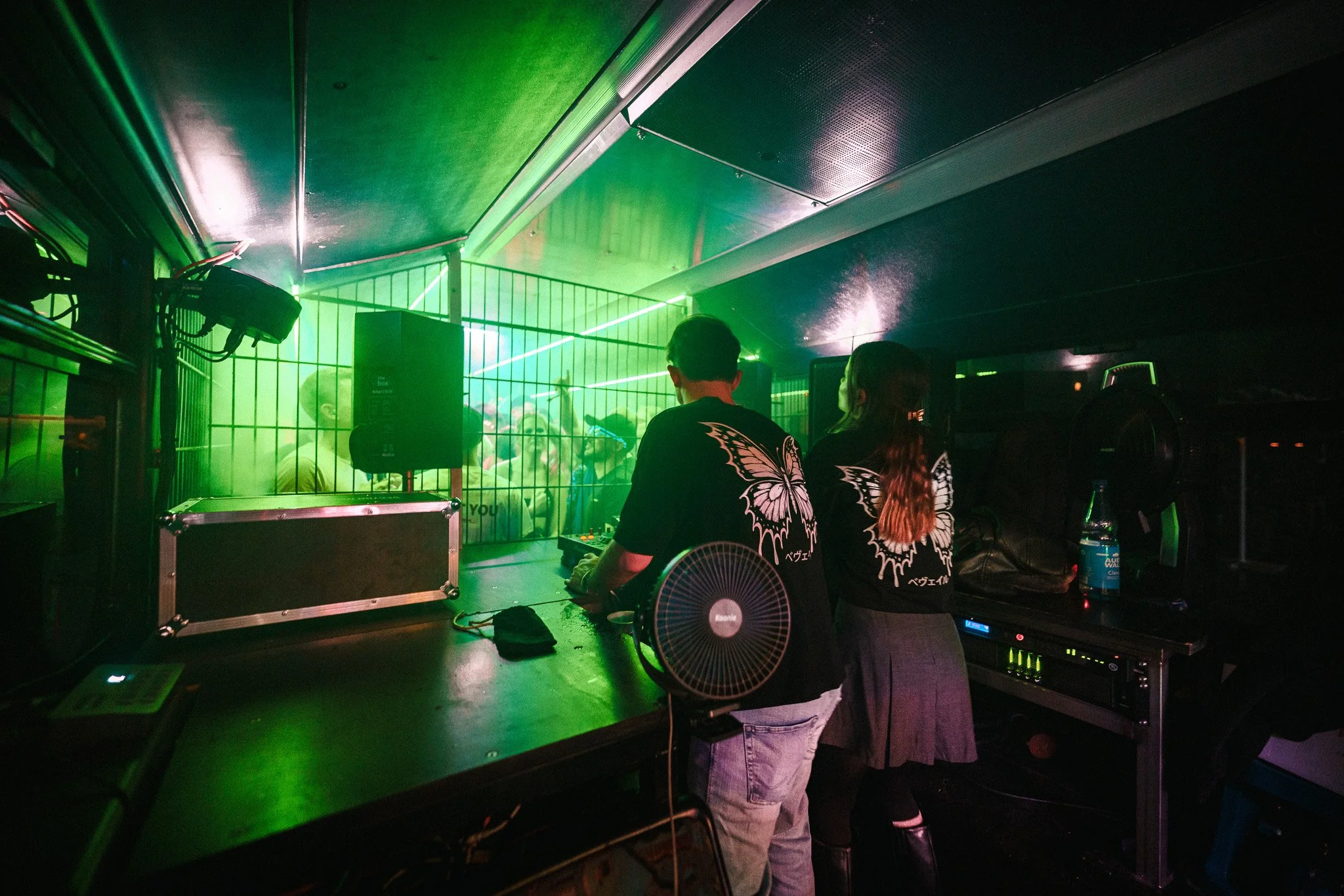 Two people DJing in a dark nightclub with a green-lit crowd behind a wire fence, wearing matching butterfly shirts.