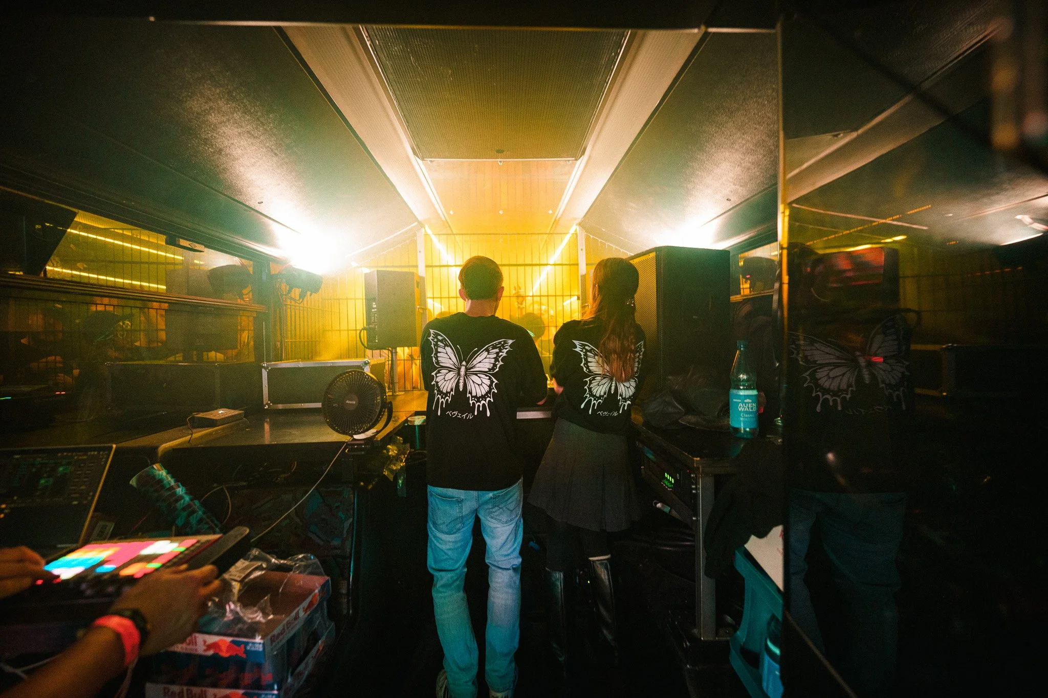 Two people with butterfly-themed clothing standing in a DJ booth at a nightclub, with yellow and green lighting, music equipment, and a large speaker in the background.