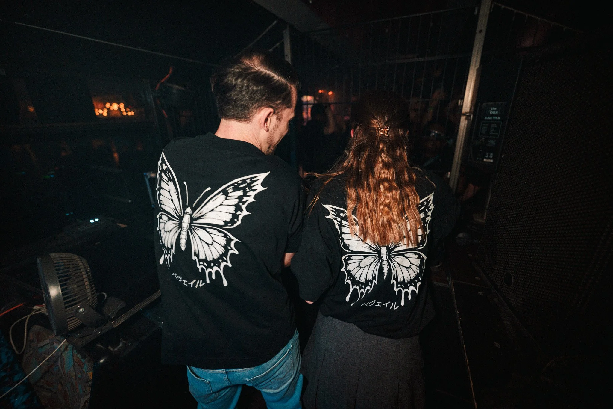 Two people with butterfly artwork on their black shirts at a nightclub or music venue, seen from behind.