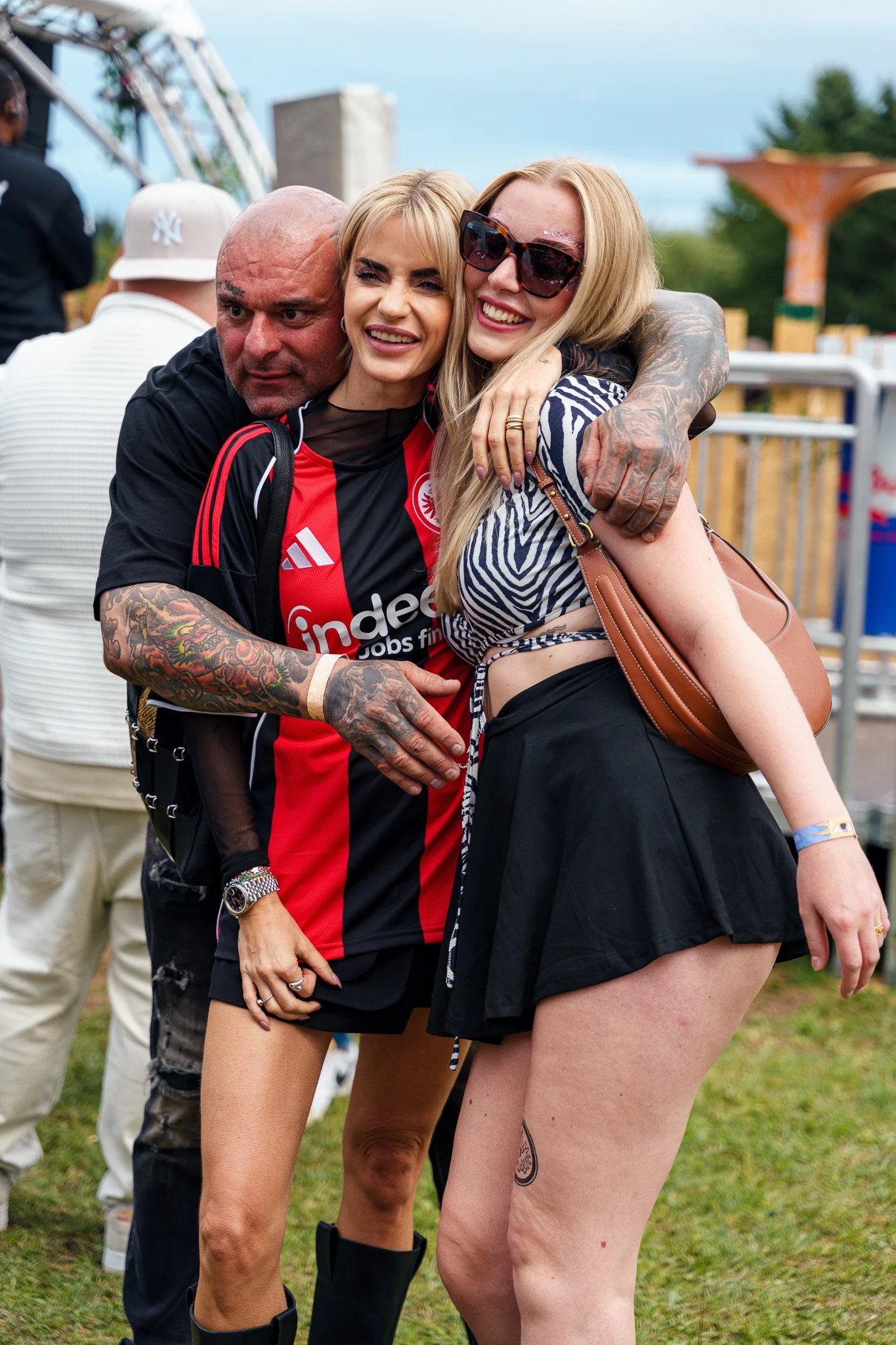 Three people hugging at an outdoor event. The person on the left is a tattooed man in a black and red sports jersey, with short hair and a serious expression. The woman in the middle has blonde hair, is smiling, and wearing a black and red sports shi