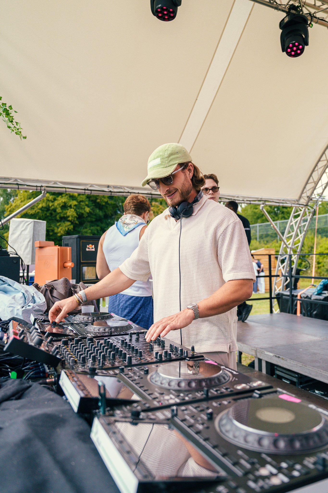 A male DJ wearing sunglasses, a beige cap, and a white short-sleeved shirt mixes music at an outdoor event under a large beige tent.