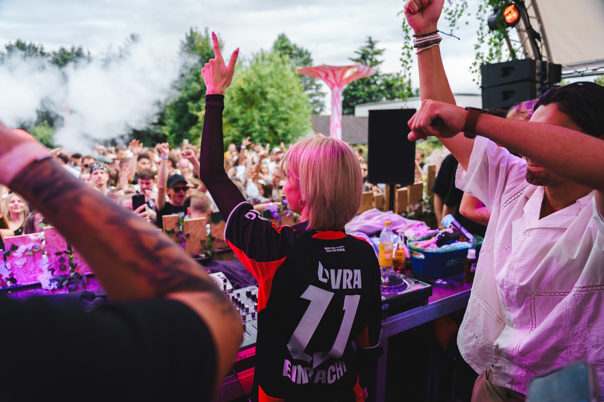 A crowded outdoor music festival with a DJ woman wearing a black and red jersey with 'LIVRA 11' on the back, raising her right hand. The crowd is cheering and dancing, with trees and stage equipment visible in the background.