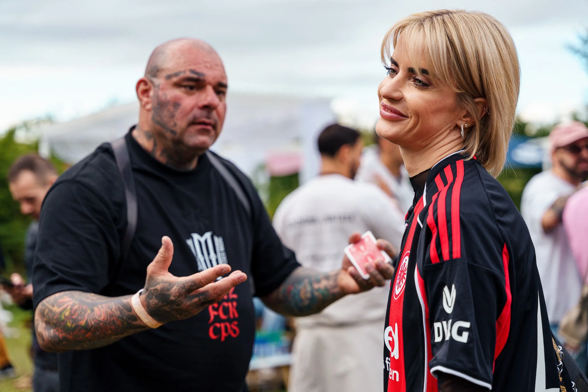 A woman in a sports jacket smiling and talking to a tattooed man holding a deck of cards at an outdoor event with people in the background.
