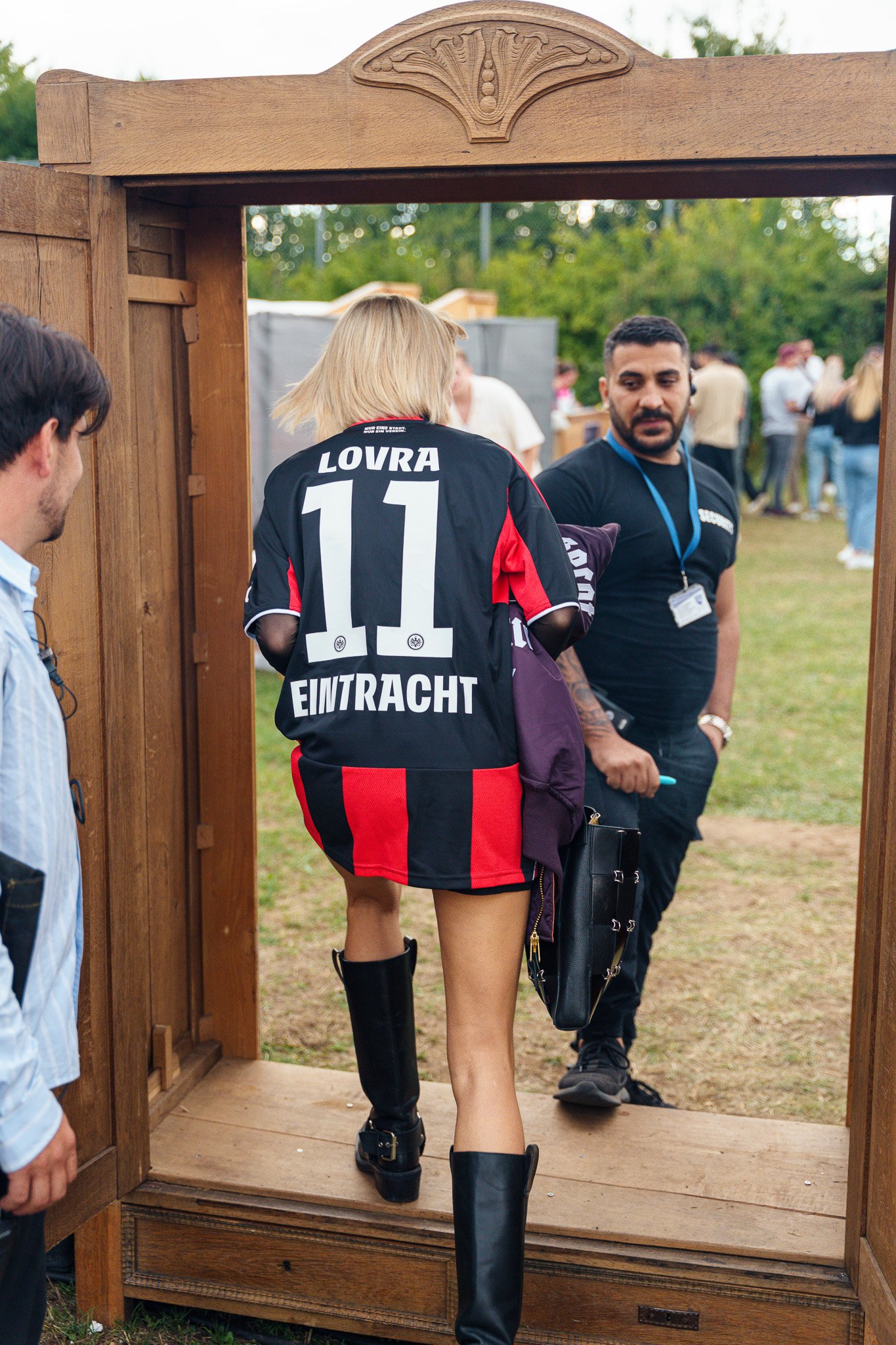 Woman with blonde hair wearing a black and red soccer jersey, number 11, with 'LOVRA' and 'EINTRACHT' on the back, walking through a wooden archway, holding a purple jacket, with onlookers in the background.